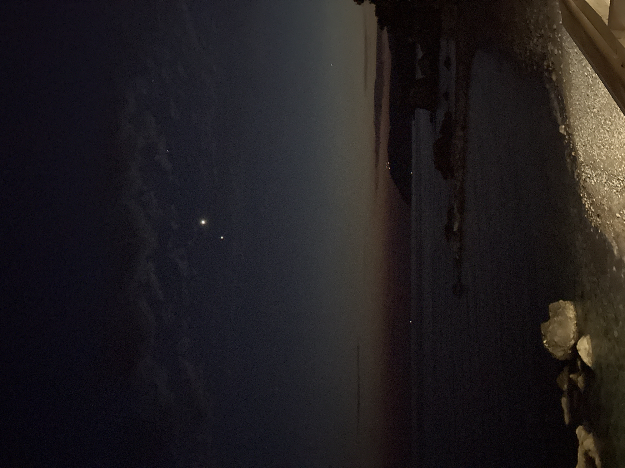 The image depicts a nighttime view of the Adriatic Sea coastline in Šušanj, Montenegro. The sky is a deep, dark blue, transitioning to a faint orange and pink gradient near the horizon, indicative of twilight or post-sunset light. Two bright celestial bodies are visible in the sky; one is higher and to the left, appearing as a distinct point of light. The second celestial body is lower and to the right, also a bright point. A thin, wispy cloud formation is present in the upper portion of the sky. The sea is dark and calm, with gentle waves lapping against a pebble beach. Several large, light-colored rocks are visible in the foreground and near the water's edge. In the distance, a silhouette of a coastal town or village with scattered lights can be seen along the hills. The overall atmosphere is serene and natural, with a hint of urban presence on the distant shoreline.