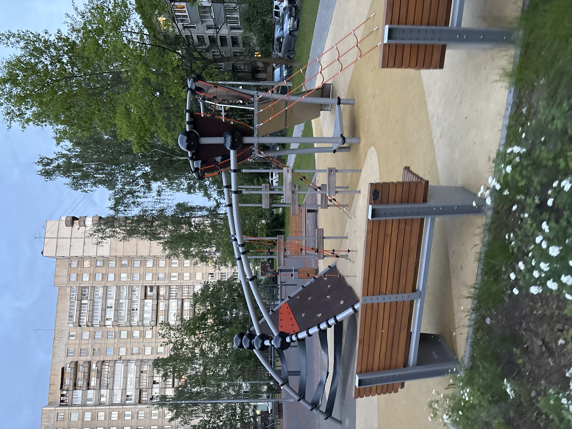 This image captures a modern, unoccupied playground structure set against a backdrop of urban residential buildings and lush trees. The playground, occupying the foreground and midground, features a contemporary design with prominent silver metal poles, black spherical connectors, and brown wooden elements. It includes climbing sections with black panels and orange ropes forming nets and bridges, suggesting various play activities. Several benches with slatted wooden seats and metal frames are integrated into the playground's design.
The ground around the play area is a mix of light yellow and tan paving, with patches of vibrant green grass adorned with small white flowers. Tall, leafy green trees frame the scene, with some obscuring parts of the background. In the background, a large, multi-story residential building, constructed from light-colored concrete, stands tall. It exhibits numerous windows and enclosed balconies, characteristic of apartment complexes found in post-Soviet architecture. A few cars are visible parked near the building.
The sky is a soft, clear blue, suggesting either late afternoon or early evening. The lighting is gentle, creating soft shadows and a peaceful atmosphere. No people are present, lending a quiet solitude to the scene. There is no visible text in the image. The overall impression is one of a well-maintained, modern urban recreational space, ready for activity.