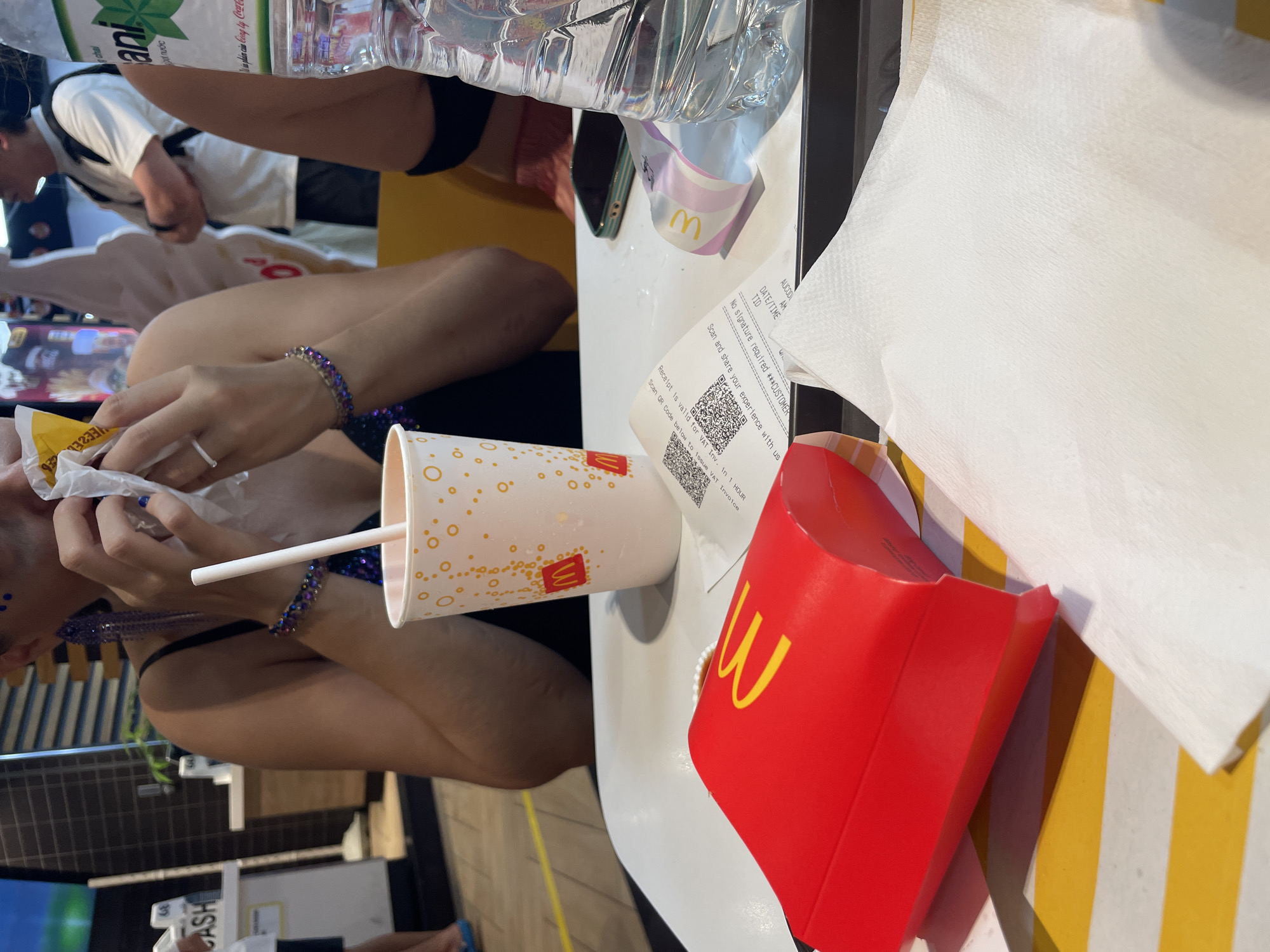 The image captures a close-up view of a table setting at a McDonald's restaurant in Thuận An, Vietnam. The focus is on a red McDonald's french fry container, an open white paper wrapper with food inside being held by a person, a white paper cup with the McDonald's logo and orange circular pattern, a plastic straw, a receipt with QR codes, a phone with a green case, and a partially visible plastic water bottle. In the background, blurred figures of people and restaurant decor are visible, including a counter with a screen that displays "CASH". The overall scene suggests a meal in progress or just concluded, with the items arranged on a white table surface.