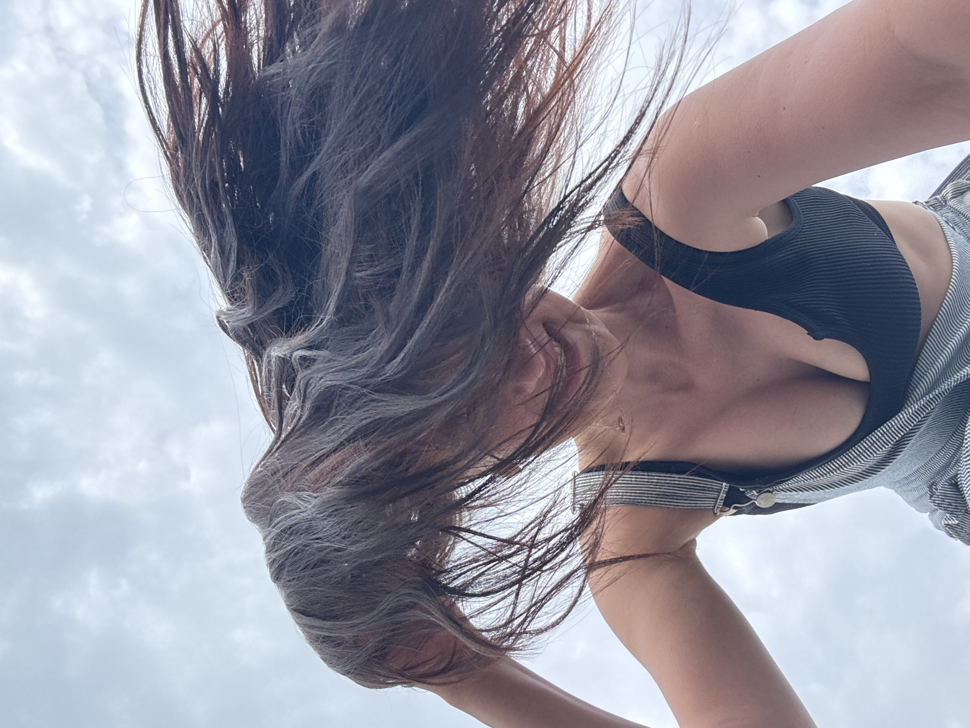 A low-angle shot captures a woman against a cloudy sky. Her dark hair is tossed, partially obscuring her face. She is wearing a black sports bra under striped overalls. The location is not discernible as Sochi, Russia, or any other specific location.