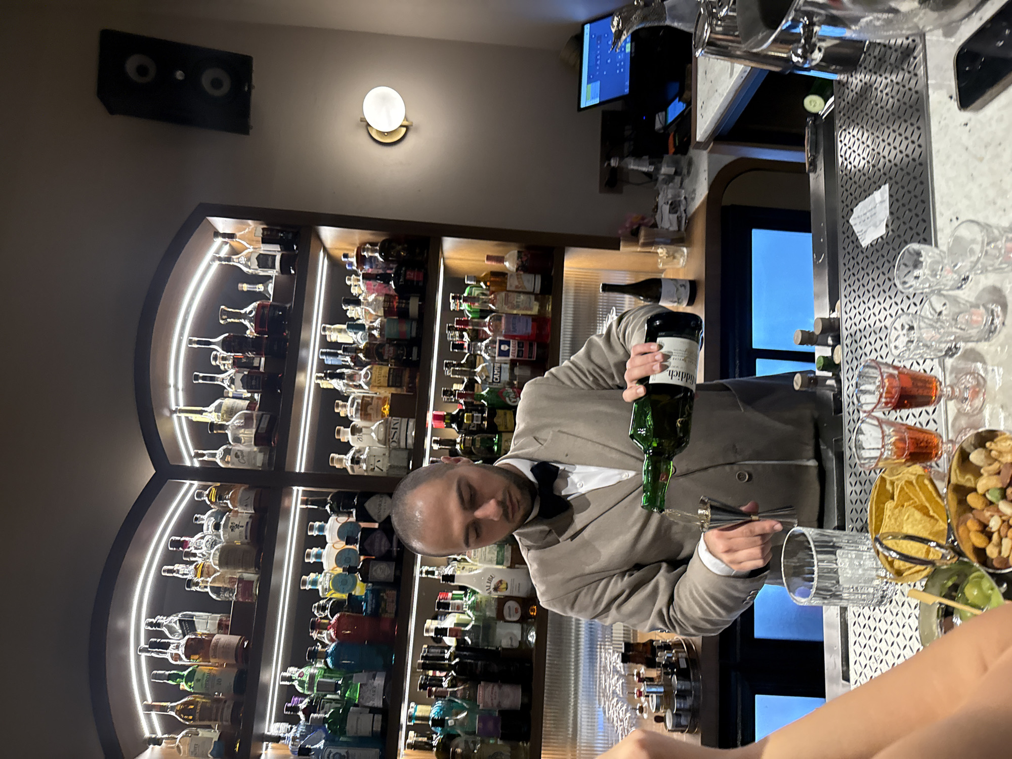 A bartender in a light beige suit and black bow tie is meticulously pouring a dark green bottle of liquid (possibly alcohol) into a jigger over a patterned glass. The bar is well-stocked, as evidenced by the liquor shelves holding numerous bottles in the background. The shelves are illuminated by arching LED strip lights, and a light fixture is present on the wall. A flat screen is visible near the counter, indicating a modern bar setup. The counter is lined with various items including nuts and tortillas. The scene appears to be at a bar, and the bartender is likely preparing a drink for a customer. Given the mention of Yerevan, Armenia, in the instructions, it can be inferred that this is a bar in that city.