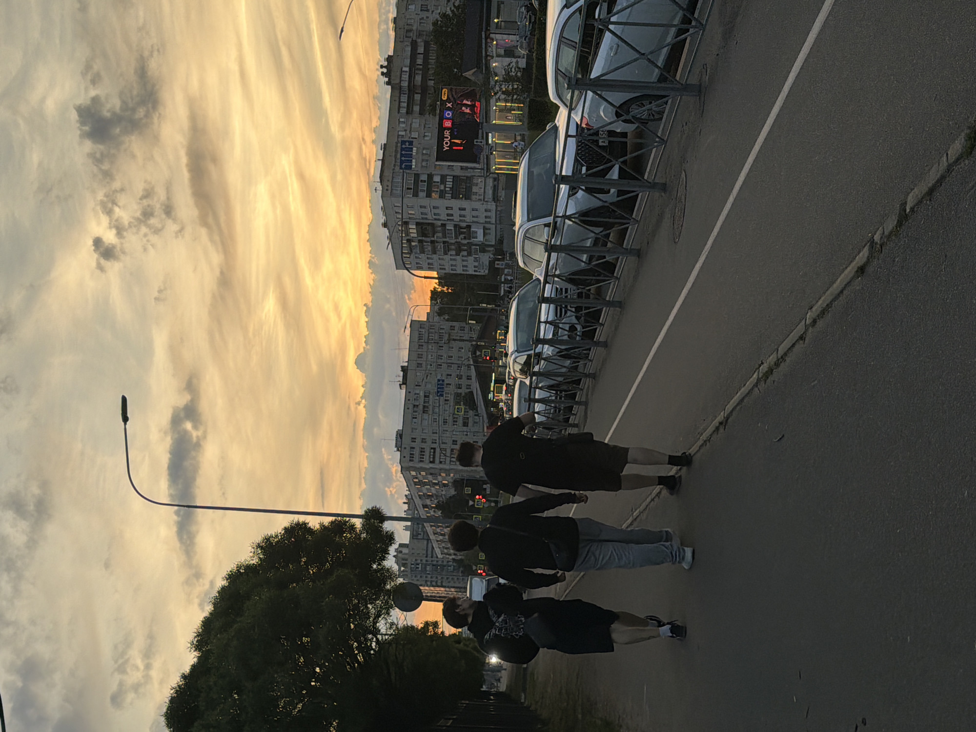 Three young men walk away from the camera down a paved sidewalk in an urban setting, likely Russia, during sunset. The sky is filled with dramatic, colorful clouds in hues of orange, yellow, and gray, illuminated by the setting sun. A street lamp with a bent pole is visible on the left side of the frame, near a dense green tree. On the right side of the sidewalk, a metal fence separates the pedestrian area from a street where several white SUVs are parked. Behind the street, multi-story apartment buildings line the horizon, with some illuminated windows visible. A large digital billboard displaying the text "YOUR BOX" is prominent on one of the buildings. The men are dressed casually; the one on the left wears a black hoodie and shorts, the middle man wears a black jacket and gray pants, and the man on the right wears a black t-shirt and shorts. All three appear to be walking in the same direction, with no discernible interaction between them. The overall atmosphere is one of a late evening stroll as the day transitions into night.