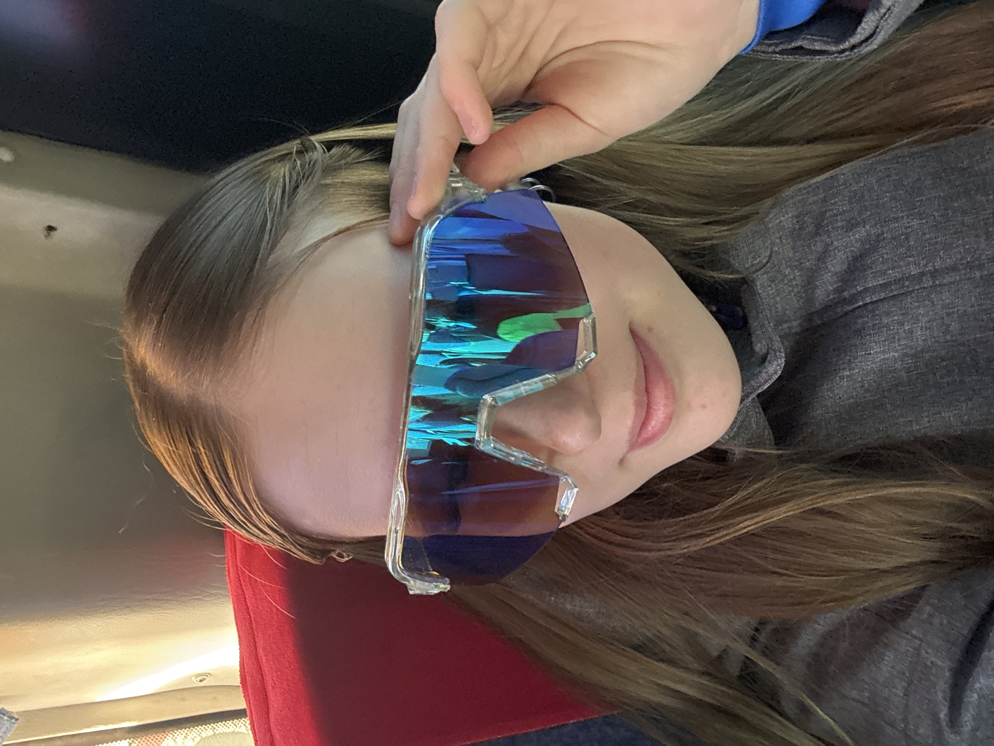 A photograph depicts a person with light brown hair, wearing blue-tinted glasses with clear frames, seemingly indoors. The person is holding the glasses with their right hand. The individual is wearing a gray jacket, and a red object is visible in the lower left corner. The location is unknown and is NOT specified as Berezniki, Russia. No other details about the environment or interactions can be determined from the image.