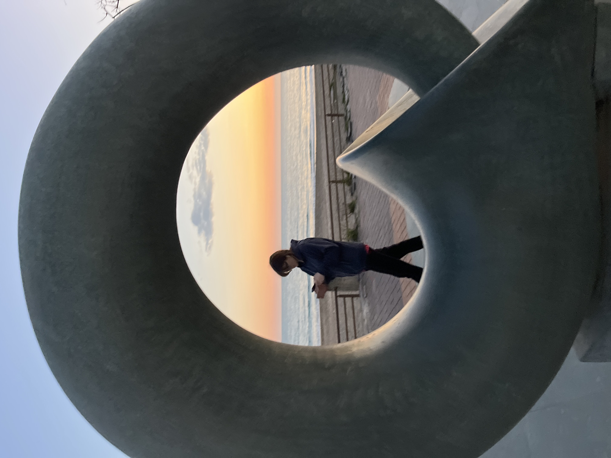 A photo taken at a low angle shows a person in dark pants and a blue jacket leaning on a railing at a beach while holding a cell phone. The person is framed by a circular sculpture that is made of gray concrete and has a complex, intersecting design. Beyond the railing is the beach, the ocean, and a colorful sunset of blue, orange, yellow, and white. The beach has a paved walkway. It is the city of Alanya, Turkey.