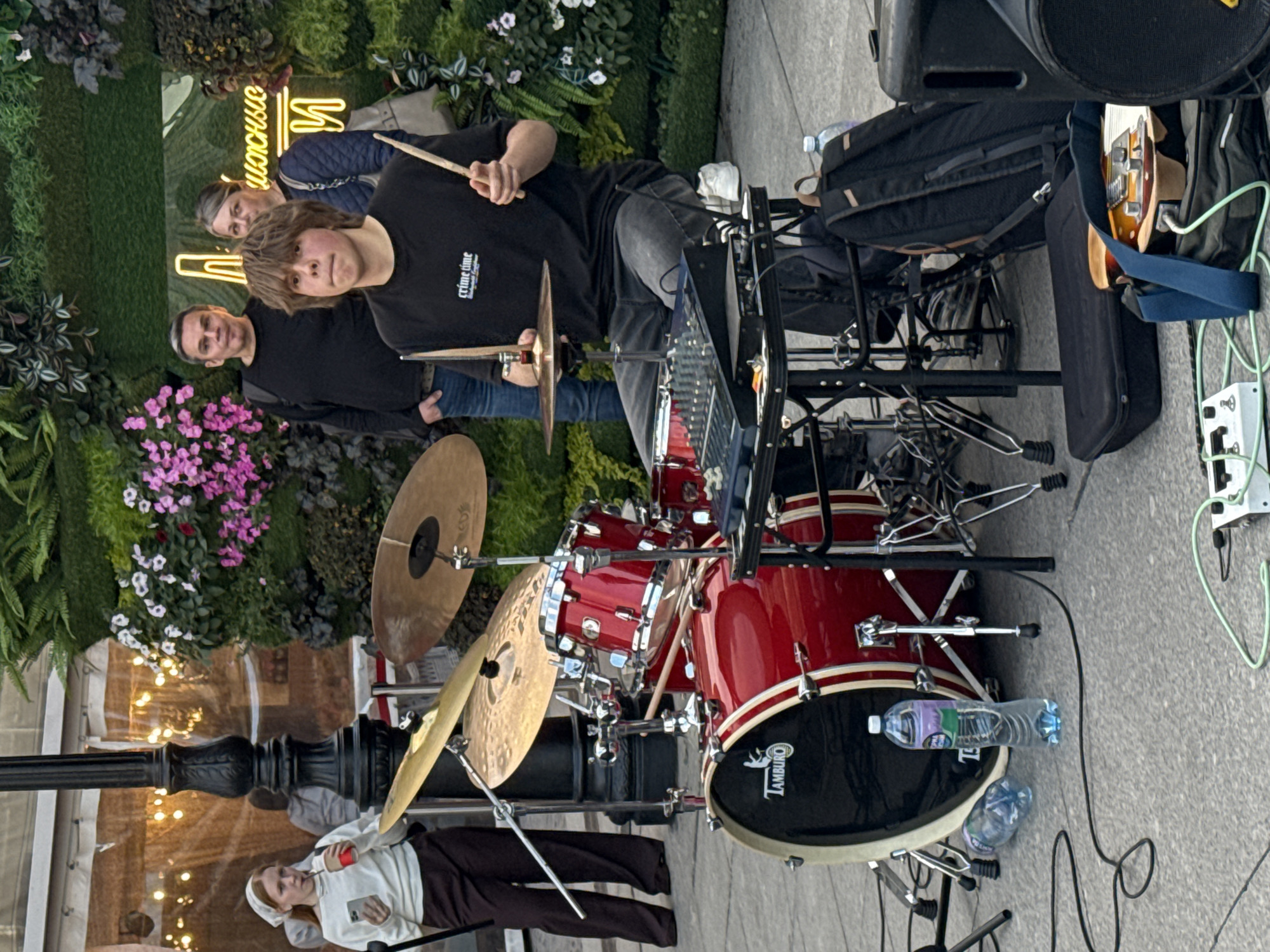 A young male is playing drums on an outdoor street in Saint Petersburg, Russia. He is wearing a black t-shirt and jeans. He is seated behind a red drum set and is holding drumsticks. A sound mixing board is on a stand in front of him. To his right, there is a black backpack. A woman and two men are standing behind him and to his left. The woman is holding a beverage and a phone. The background features a green wall with pink flowers and neon lights. The ground is paved with grey stones. There are several water bottles, cables, and speaker equipment around the drum set.