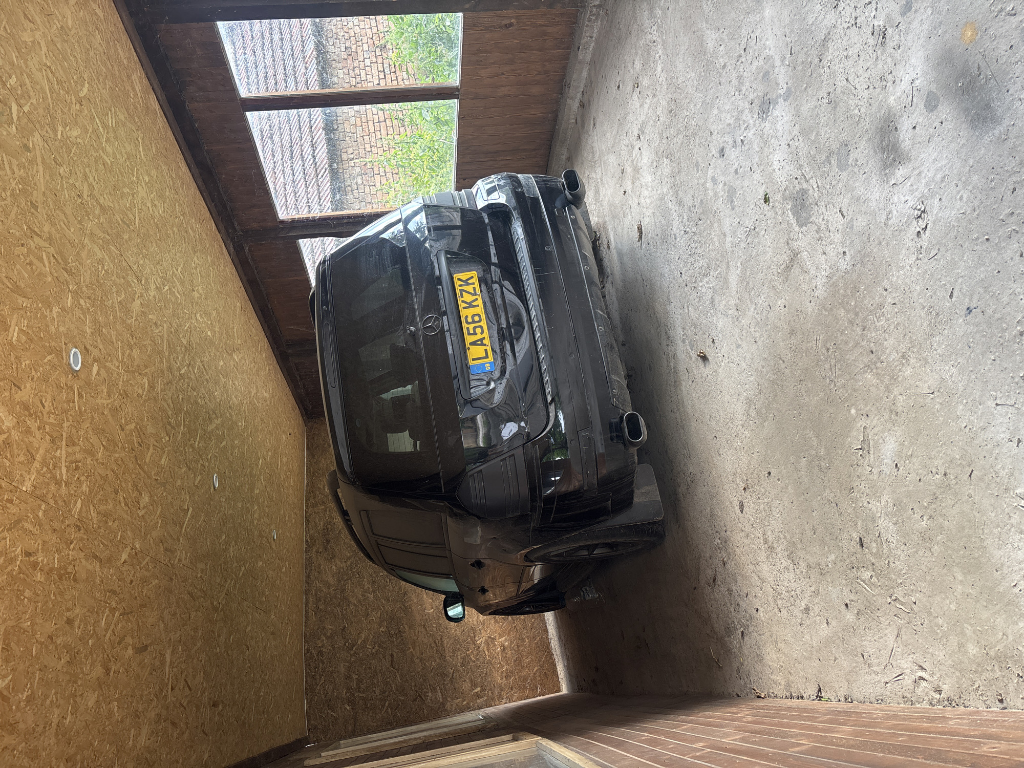 The image depicts the rear of a black Mercedes-Benz SUV parked inside a covered structure with OSB (Oriented Strand Board) walls and ceiling. The structure appears to be a makeshift garage or carport. The floor is concrete, with some debris and leaves scattered on it. Through windows on the right side of the structure, a brick wall and overgrown vegetation are visible, suggesting an outdoor environment beyond. The license plate of the vehicle is visible and reads "LA56 KZK". The lighting within the structure is dim, with two circular ceiling lights visible in the OSB ceiling. The vehicle is clean and appears to be in good condition. The location is identified as Brenitsa, Bulgaria.