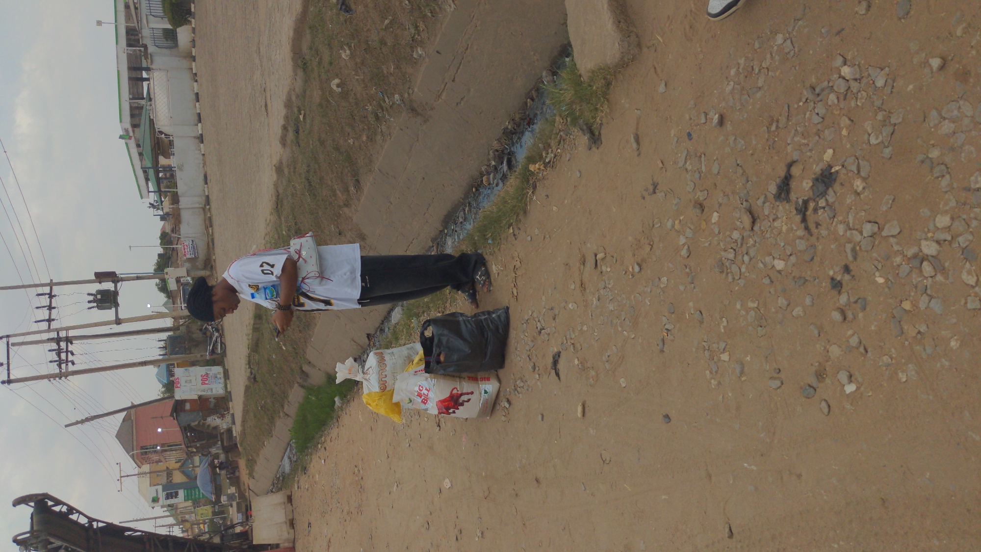 An outdoor, eye-level shot depicts a person standing on a dirt path next to a concrete drainage ditch. The person, a male, is wearing a white short-sleeved shirt with "02" printed on the sleeve, black pants or a long skirt, and a black head covering. He appears to be bending slightly forward, possibly looking at or interacting with items on the ground.
Beside him, several bags are placed on the dusty, rocky ground. These include a black plastic bag, a yellow bag, and two white sacks. One white sack clearly displays the text "BIG BULL" and features an illustration of a red bull. Another white bag has partially visible text, possibly "Omow".
To the person's left, a concrete drainage channel runs along the edge of the path, containing murky water and some sparse green grass and debris. The ground throughout the scene is uneven, a mix of reddish-brown dirt and scattered small stones.
In the background, there's a street lined with buildings and numerous utility poles with electrical wires crisscrossing the sky. Some signs are visible on buildings, one partially reading "POS". The sky is overcast, suggesting daytime. The overall setting appears to be a busy, urban or semi-urban street in Ado, Nigeria. The scene shows aspects of daily life and infrastructure.