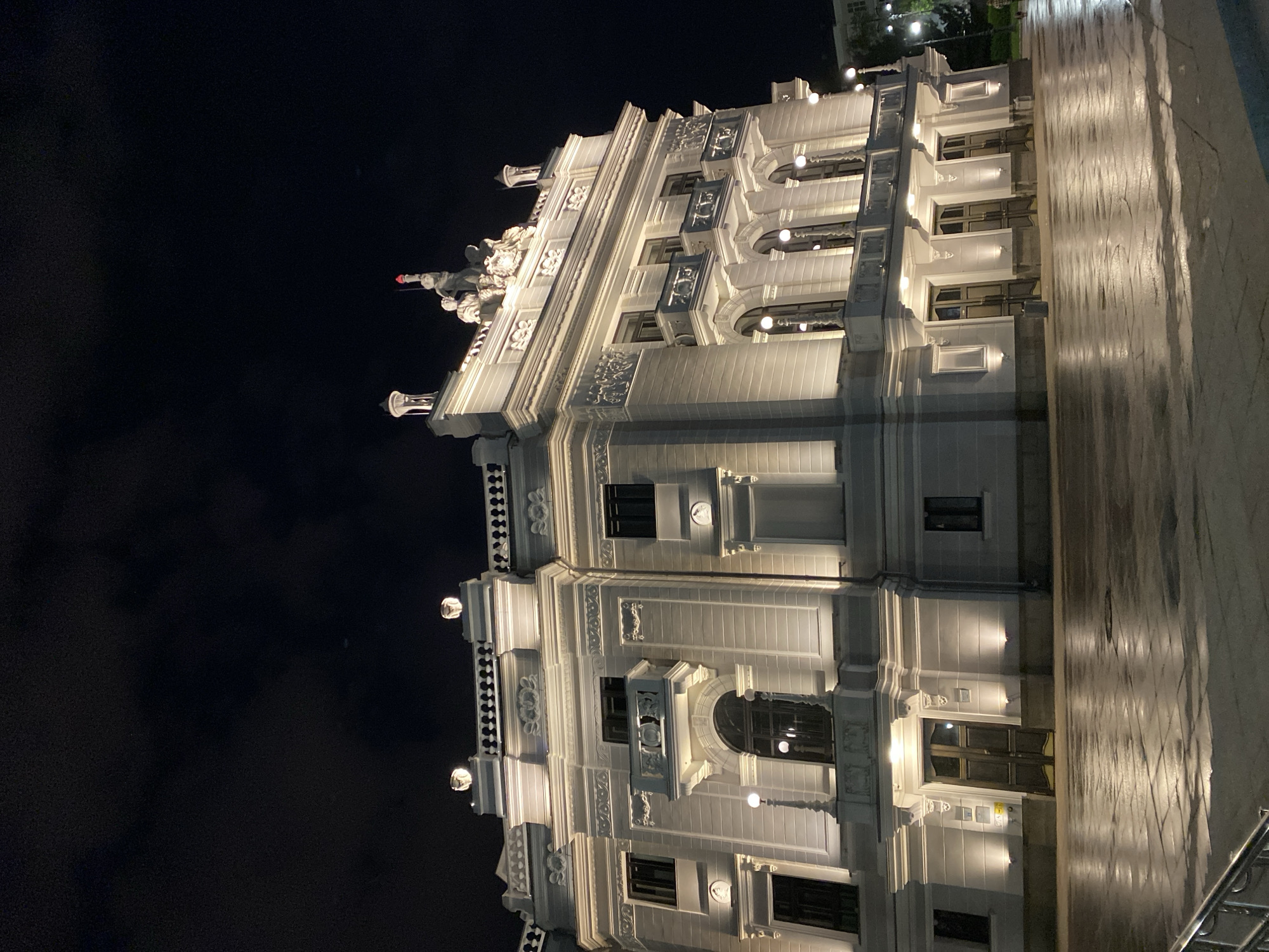 The photograph depicts the illuminated facade of a building at night. The building is white and features classical architectural details such as ornate moldings, arched windows, and decorative embellishments. Streetlights are mounted on the building. The foreground shows a sidewalk surface. The sky is dark. The image is of the building located on Vaynera Street in Yekaterinburg, Russia.