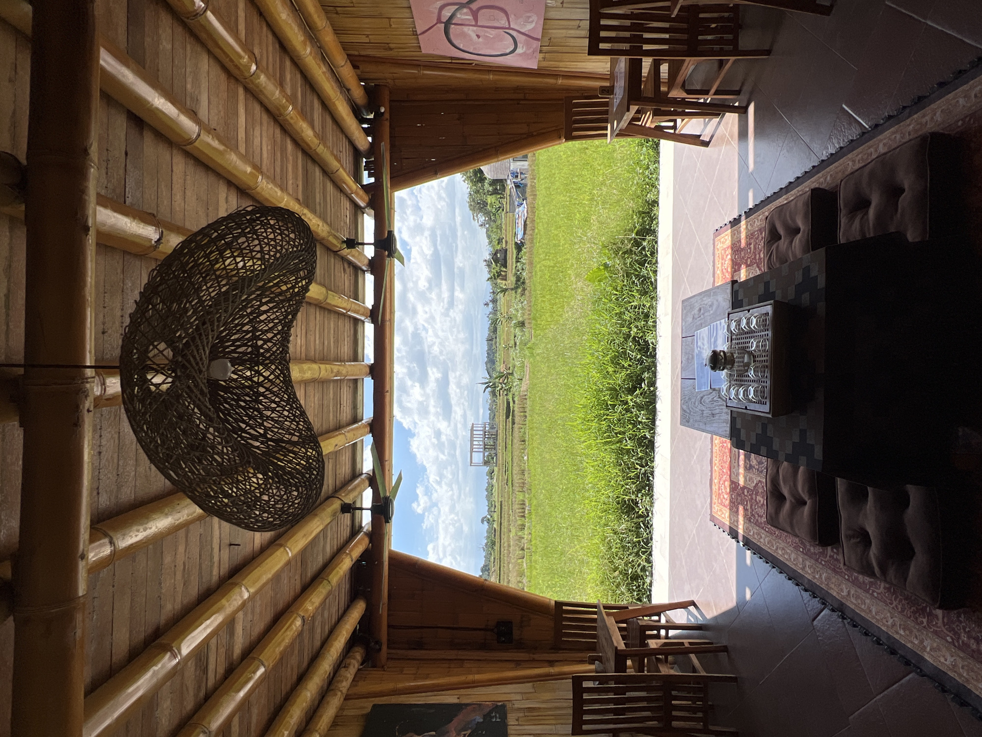 The image depicts an indoor space with a view of a lush, green field in Ubud District, Indonesia. The interior structure is primarily constructed of bamboo, forming walls and a ceiling. A large, woven lamp hangs from the ceiling, and simple wooden furniture, including chairs and a low table with glassware, is arranged within the space. Cushions are placed along the floor near the table, which sits on a rug. The field outside is filled with tall grass and has a couple of structures visible in the distance under a sky with scattered clouds.