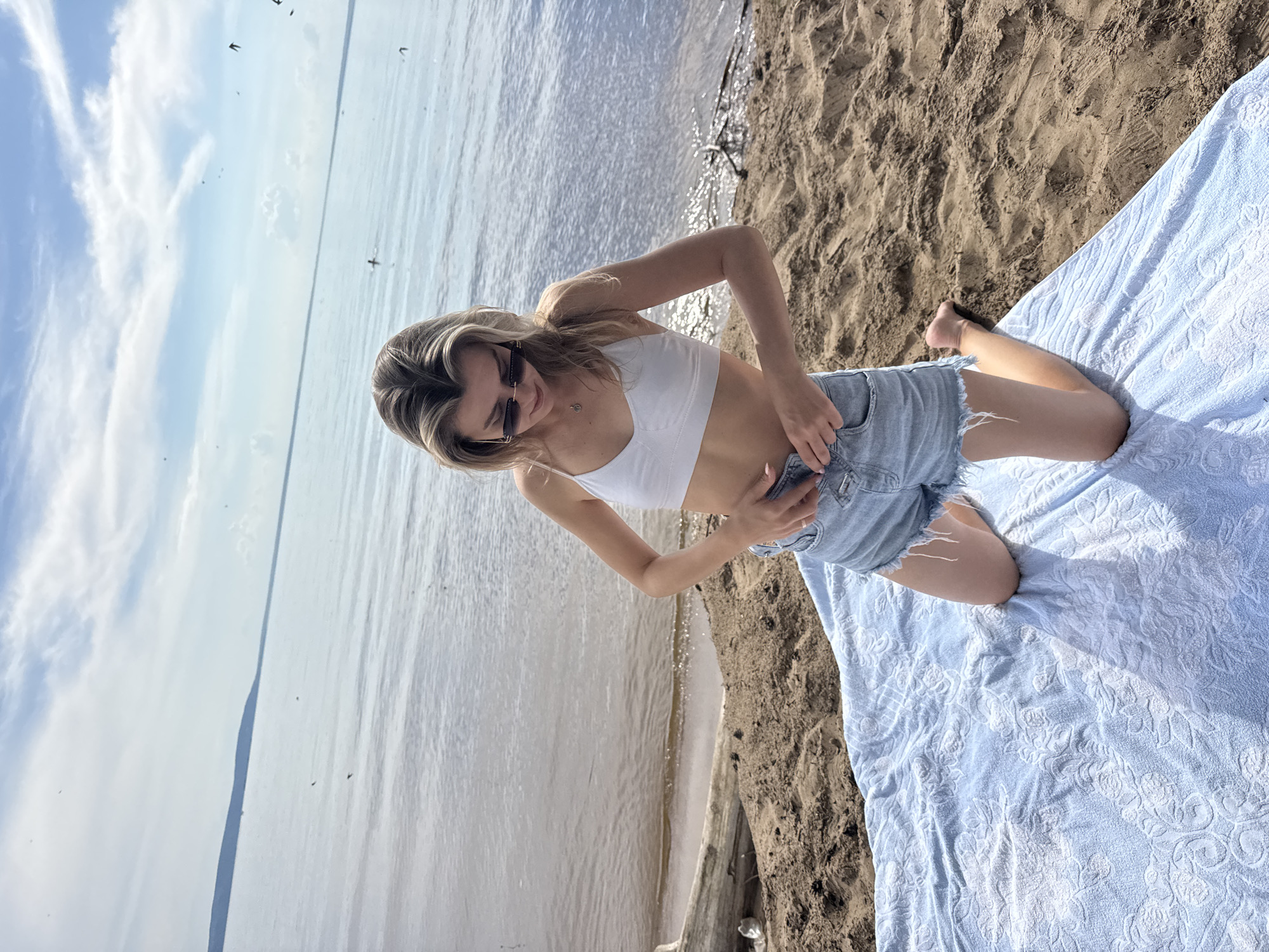 A young Caucasian woman with blonde hair, wearing a white bikini top and denim shorts, kneels on a light blue towel spread on a sandy beach. She is looking down and adjusting her shorts. Her sunglasses are perched on her nose. The background shows a large body of water under a cloudy blue sky. There are some birds in the sky and the water. A piece of driftwood lies on the sand to the left of the woman.