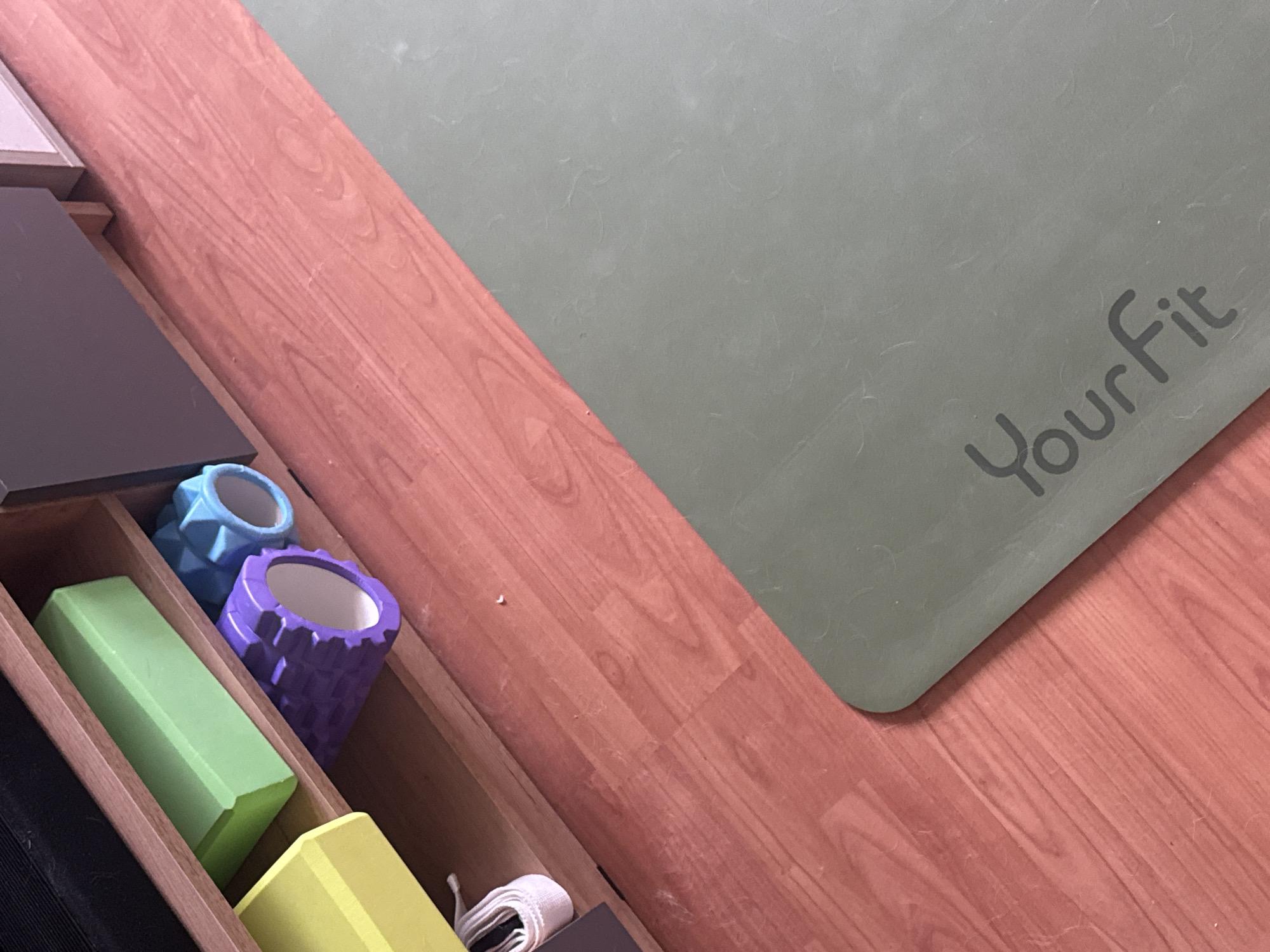 A yoga mat with the "YourFit" logo is laid out on a wooden floor. To the left of the mat, several yoga accessories are stored in a wooden organizer. These include a green foam block, a blue textured massage roller, and a purple textured massage roller. There is also a yellow foam block and a white yoga strap visible in the organizer. The lighting suggests an indoor setting, possibly a home or studio. The scene appears to be still and organized, with no activity actively taking place. The image is taken from a high-angle perspective, looking down at the floor and the yoga equipment. The wooden floor has a reddish-brown hue and a visible wood grain pattern. The yoga mat is a muted green color. The overall impression is one of preparedness for a yoga or fitness session.