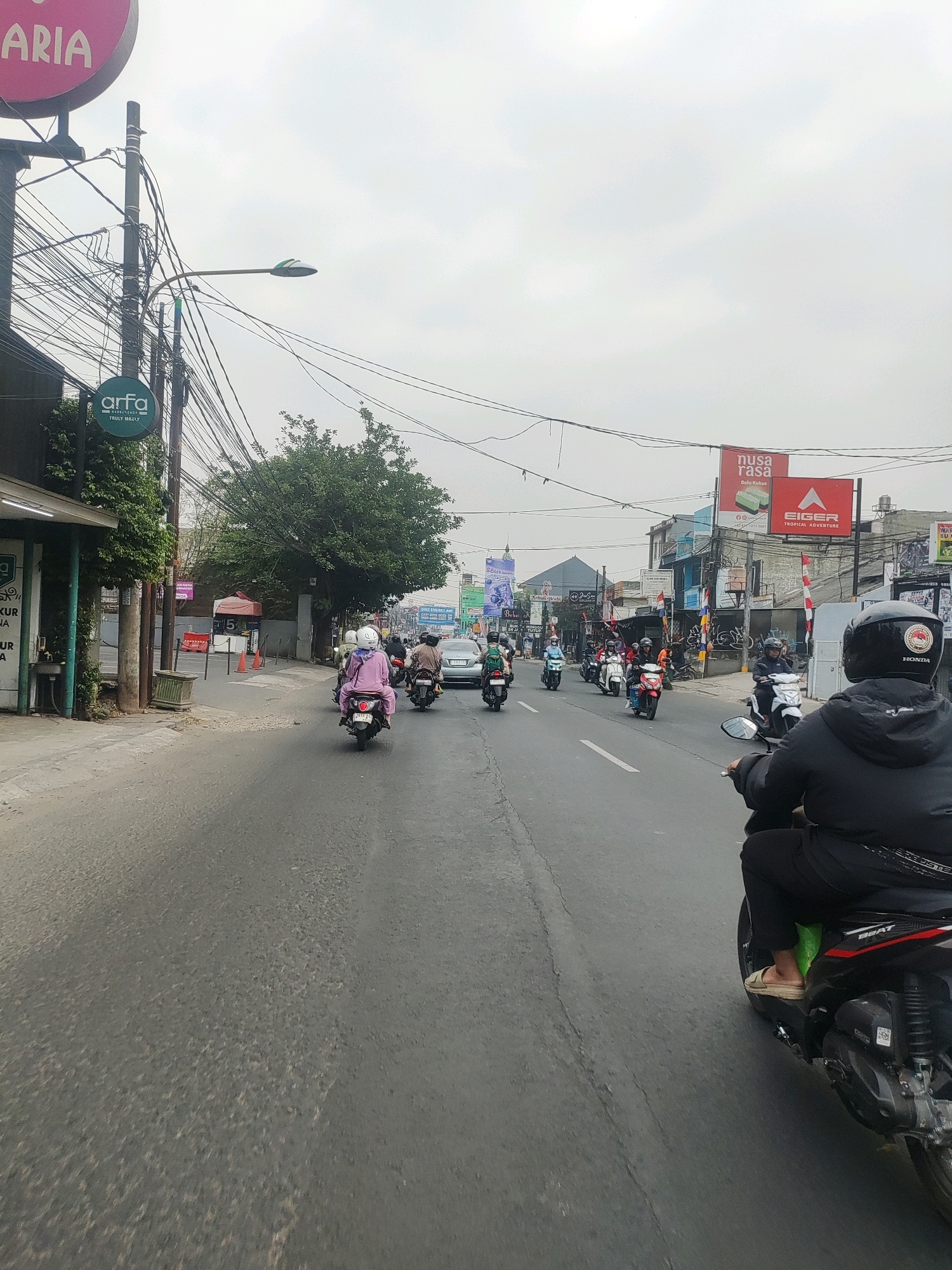 This is a photograph taken from a moving vehicle, likely a motorcycle, on a street in Bekasi, Indonesia. The street is paved and appears to be moderately busy with vehicular traffic, primarily motorcycles. On the left side of the road, there is a sidewalk, a building with a green awning, and a pole with numerous electrical wires strung across it. A circular pink sign with the word "ARIA" is visible at the top left. Further down the street, there are more buildings, trees, and various signs, including "Arfa" with text below it, and banners with text and logos. On the right side of the street, there are also buildings, signs, and more traffic. Prominent signs include "nusa rasa" and "EIGER TROPICAL ADVENTURE". A person in a black jacket and helmet is riding a motorcycle in the foreground, facing away from the camera. Other motorcyclists and a silver car are visible further down the street. The sky is overcast.