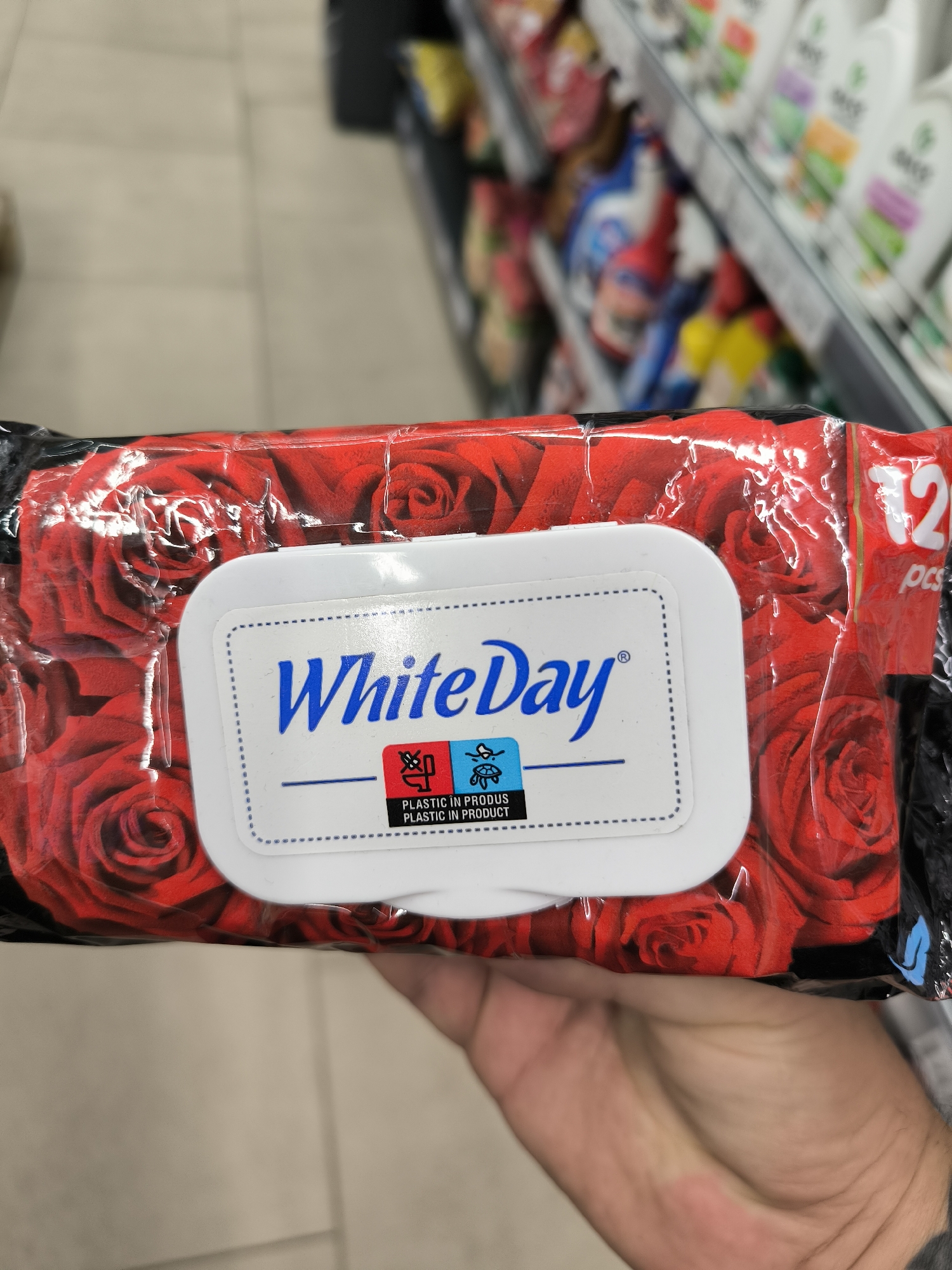 A consumer holds a package of "White Day" wet wipes with a red rose design. The wipes are packaged in clear plastic, revealing the floral pattern underneath. A white label on the package displays the brand name in blue, along with two small icons. Below the icons, text reads "PLASTIC IN PRODUS" and "PLASTIC IN PRODUCT." The background appears to be a retail environment, with blurred shelves of products visible. The floor tiles suggest an indoor setting. The lighting is even and appears to be artificial, typical of a store.