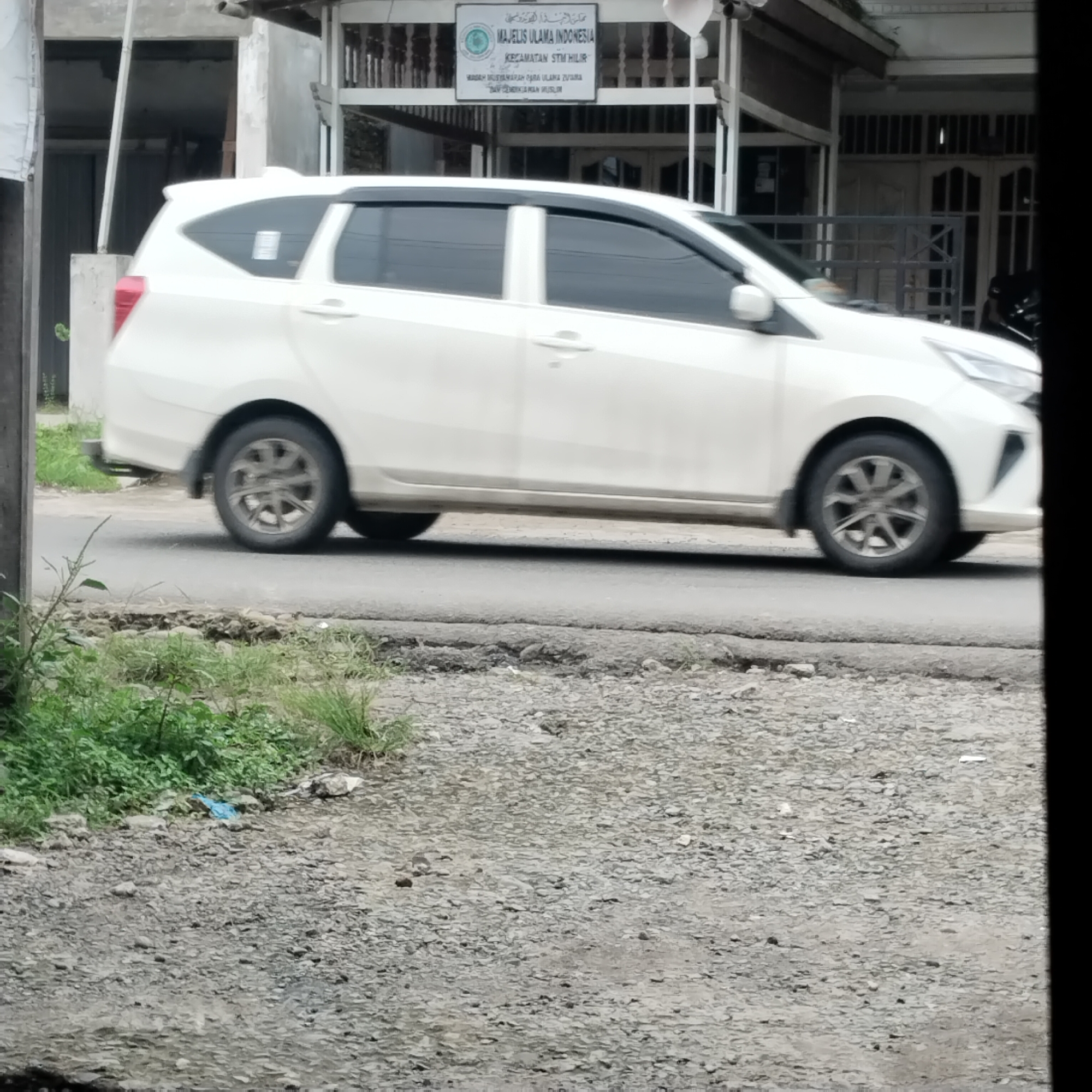 A white MPV is driving down a paved road. The car is captured from the side, in motion. To the left of the road, there is a patch of green grass and some rocks. In the background, there is a building with a sign that reads "MAJELIS ULAMA INDONESIA KECAMATAN STM HILIR". The building appears to be a mosque or a religious institution. The overall scene is a snapshot of everyday life in a suburban or rural area of Indonesia, possibly Talun Kenas.