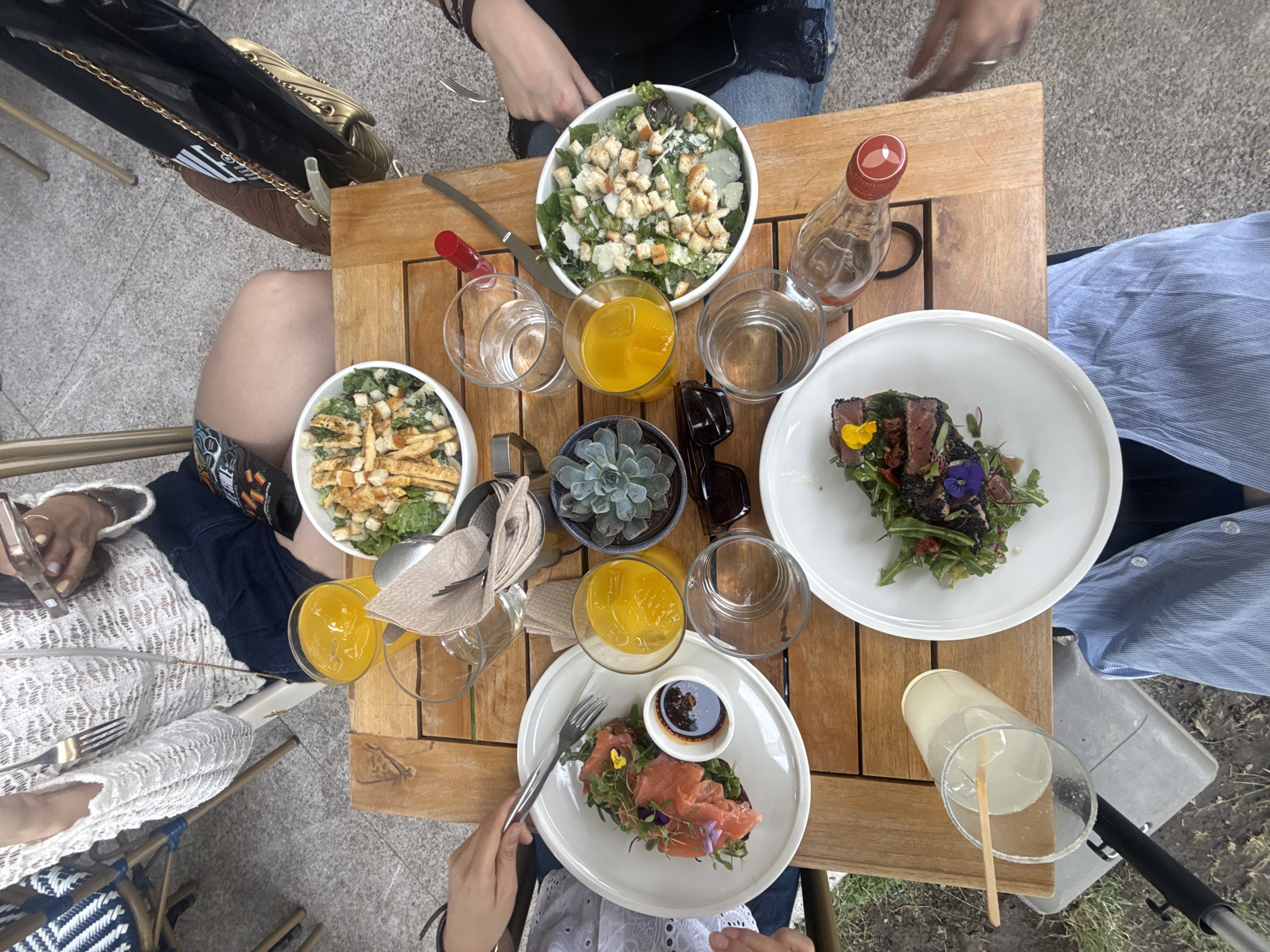The image shows a top-down view of a wooden table set with food and drinks, with sections of four people visible around the table. There are two bowls of salads with croutons, two white plates with garnished food, three glasses of orange juice, and three glasses of water. A small potted succulent plant and a pair of sunglasses sit in the center of the table. One person is holding a fork, appearing to be eating. The environment suggests an outdoor setting, possibly a patio or cafe in Mexico City, Mexico.