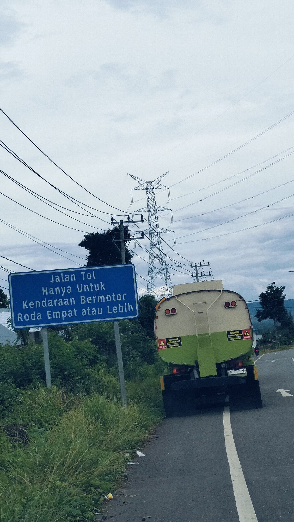 The image captures an outdoor road scene in Payakeureuleh, Indonesia, under an overcast, cloudy sky. A two-lane asphalt road with a white solid line on its right edge gently curves to the right, leading into the distance. The left side of the road is bordered by abundant green grass, bushes, and trees.
A prominent blue rectangular road sign stands on the left, partially obscured by a utility pole and vegetation. The white text on the sign, in Indonesian, reads: "Jalan Tol" (Toll Road), "Hanya Untuk" (Only For), "Kendaraan Bermotor" (Motor Vehicles), and "Roda Empat atau Lebih" (Four Wheels or More), indicating restrictions for the toll road ahead.
In the foreground, a light green and beige (or off-white) tanker truck is seen from the rear, driving on the road. The truck has two round red taillights and two yellow triangular warning stickers on its back, each bearing the text "KENDARAAN LEBAR & PANJANG" (Wide & Long Vehicle) and "LONG VEHICLE" below it. Its license plate, "BL 8293 WU," is clearly visible.
Numerous overhead power lines crisscross the cloudy sky, connected to a large, metallic lattice-style electricity transmission tower (pylon) in the mid-ground. Another smaller utility pole stands near the blue sign. The background features more trees and distant hills, suggesting a rural or semi-urban setting. The soft, diffused light indicates an overcast day. Some small pieces of white and yellow trash are visible on the road shoulder next to the grass.