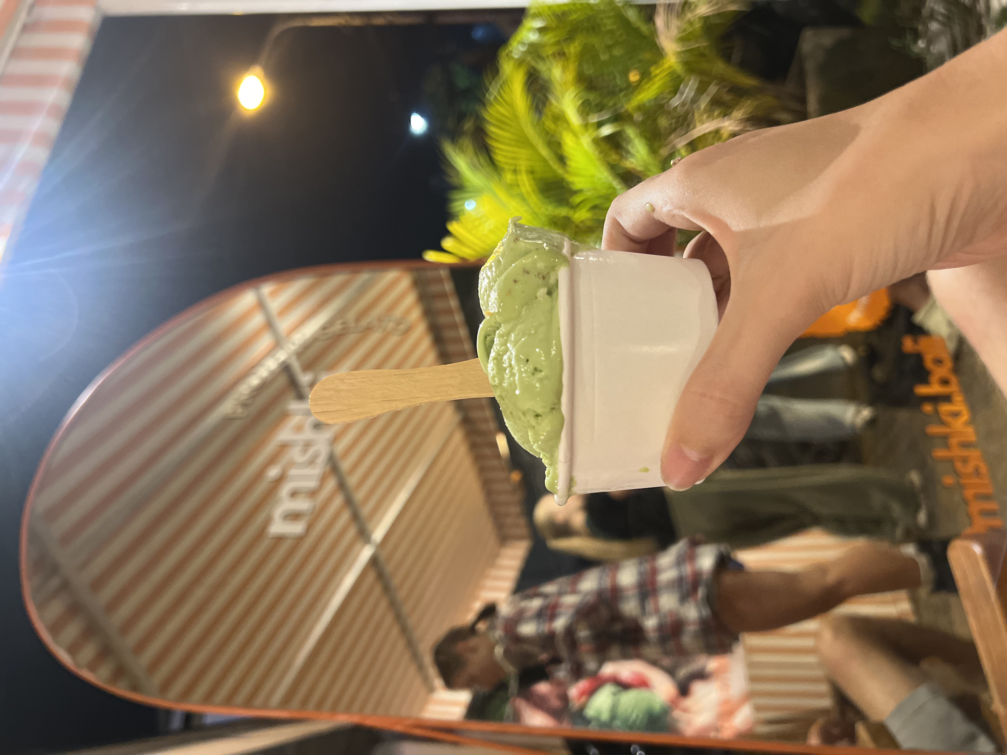 This nighttime photograph, taken in North Kuta, Indonesia, features a close-up of a hand holding a small white cup filled with green ice cream or gelato, into which a wooden spoon is inserted. The ice cream appears to be pistachio or matcha-flavored, with visible specks.
The background is dominated by a large, oval-shaped reflective surface, likely a mirror, which displays a reversed reflection of the surroundings. Partially visible text on this surface includes "mishka.bo", "mishka", and "GELATO", suggesting a business name and product offering. The reflection reveals the interior or exterior of an eatery with distinctive orange and white striped walls.
Reflected figures of people can be seen, though blurred. One individual wearing a plaid shirt (red, white, and blue/grey) is partially visible, seated within the reflected space. Further back, additional indistinct figures contribute to the scene of a bustling establishment. Overhead, a bright, yellow street lamp or bulb casts light against the dark night sky. Lush green foliage, possibly palm leaves, is also visible in the upper background, hinting at a tropical setting.
The image captures a moment of enjoying a cold dessert at what appears to be a casual outdoor or semi-outdoor eatery during the evening.
