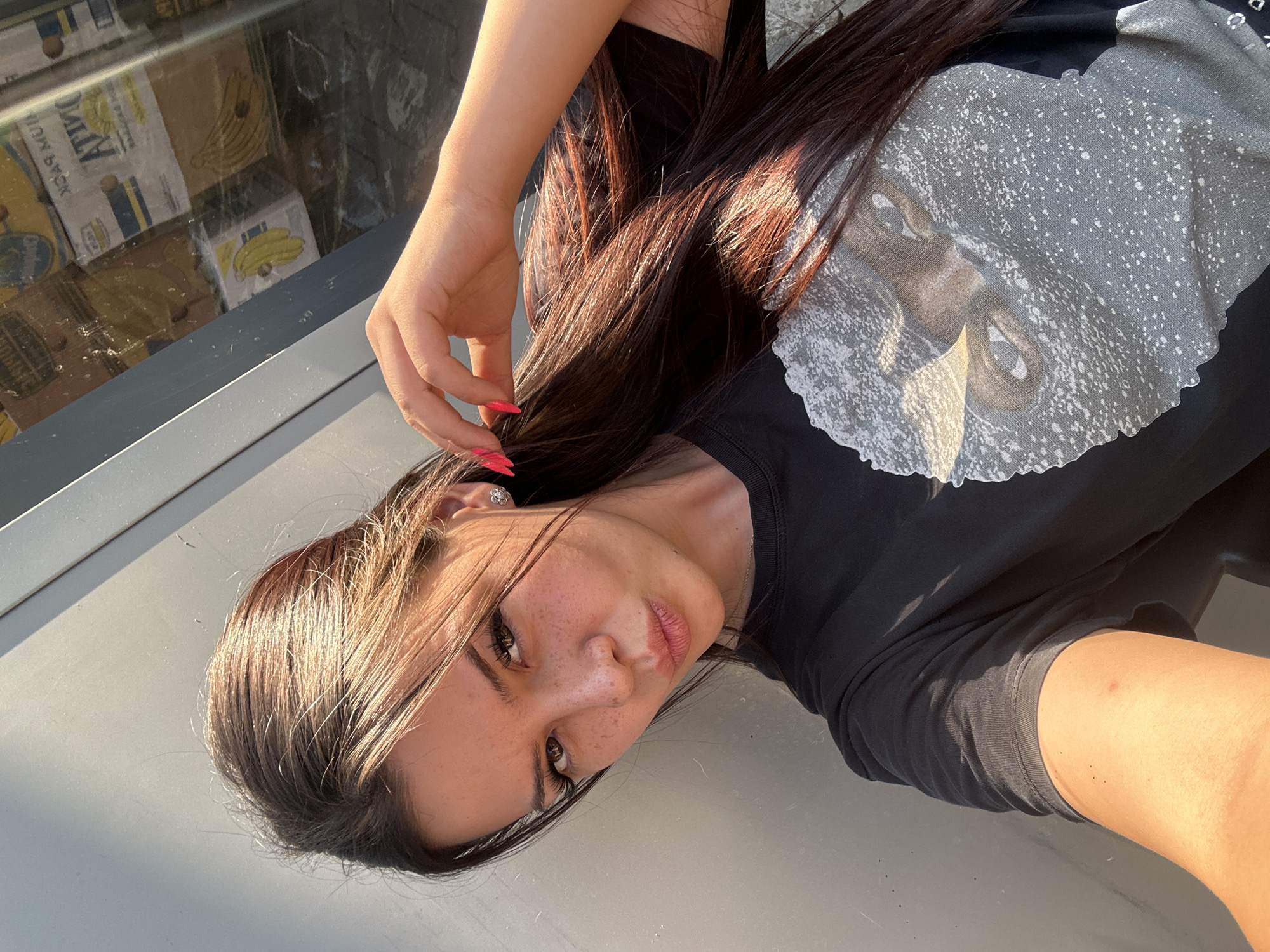 The image shows a young woman taking a selfie outdoors. She is wearing a black t-shirt with a graphic print of a face. The woman has long, dark hair and is wearing makeup, including false eyelashes and bright pink nail polish. Her hand is touching her hair. She is looking at the camera with her lips pursed. Behind her, there is a reflective surface, possibly a window, showing stacks of boxes, some of which have images of bananas. The lighting suggests it is daytime. The scene appears to be in an urban setting, possibly near a shop or warehouse. There is no indication of the location being Sliven, Bulgaria.