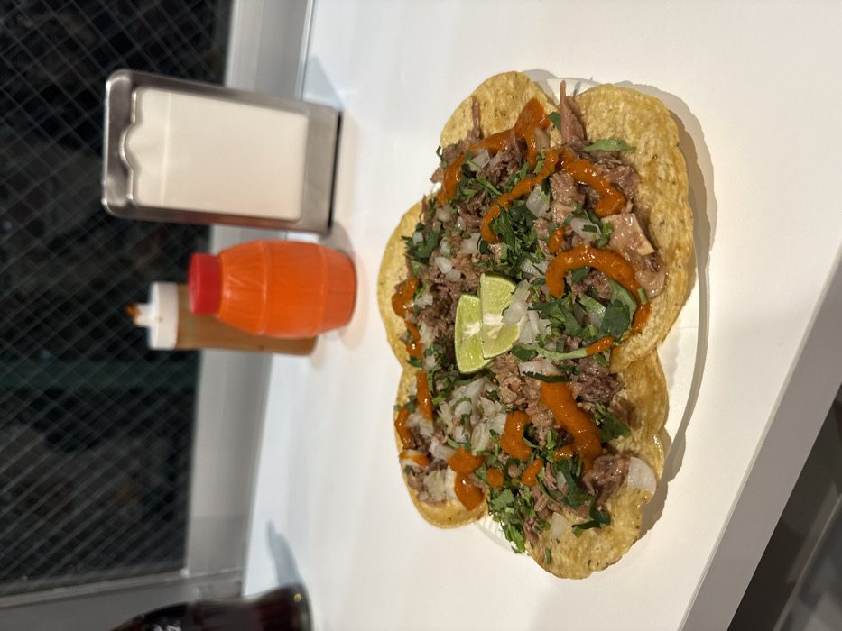A white paper plate holding five fully dressed tacos is centered on a clean, white reflective counter. Each taco features a golden-brown tortilla piled with finely shredded dark meat, topped with diced white onions, chopped green cilantro, and drizzled with a vibrant orange-red sauce. Two lime wedges are positioned centrally on the topmost tacos. To the left, a portion of a dark glass bottle is visible. Behind the tacos, from left to right, are a tall, slender condiment bottle with a light-brown sauce and white cap, an orange barrel-shaped condiment bottle with a red cap, and a silver-colored metal napkin dispenser containing white paper napkins. The background reveals a dark window with a grid pattern, suggesting an indoor setting with an obscured exterior or nighttime view. The scene is located in Shibuya, Japan.