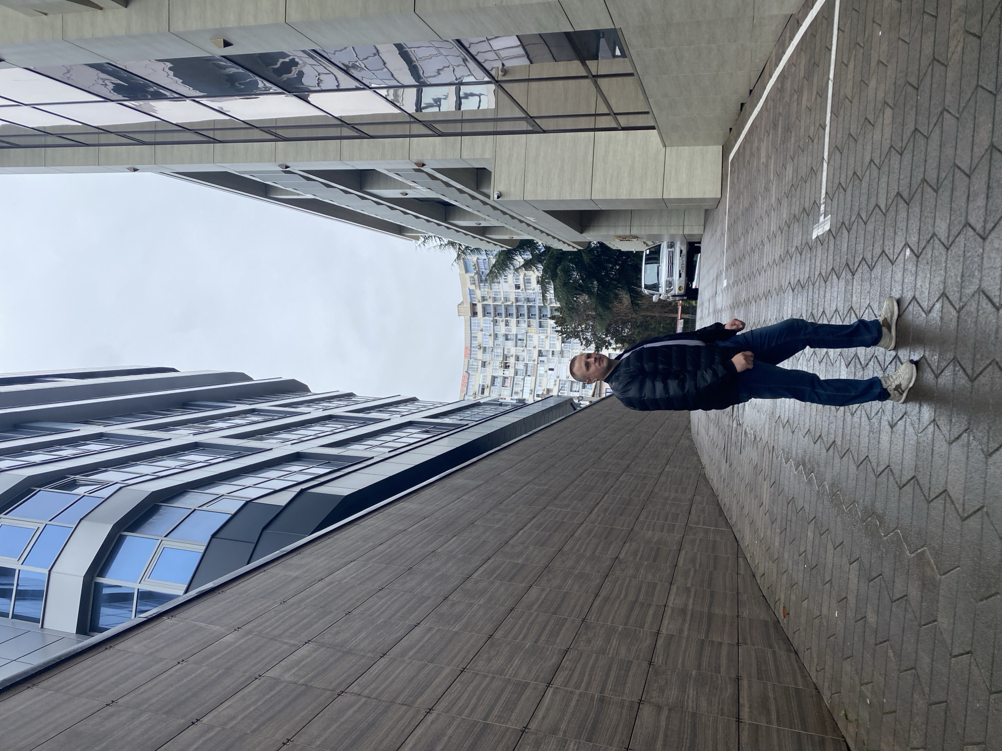 A man is standing on a paved walkway, sandwiched between two modern buildings. The pavement has a zigzag pattern and a white painted line denotes a parking space. The building on the left is tall with a mix of glass and brown tiled panels. The building on the right is a lighter grey color with reflective glass panels. In the distance, another large building is visible with many windows and balconies. The sky overhead is overcast. The man wears a black jacket and jeans. The location appears to be Sochi, Russia due to the architecture and style of the building in the background.