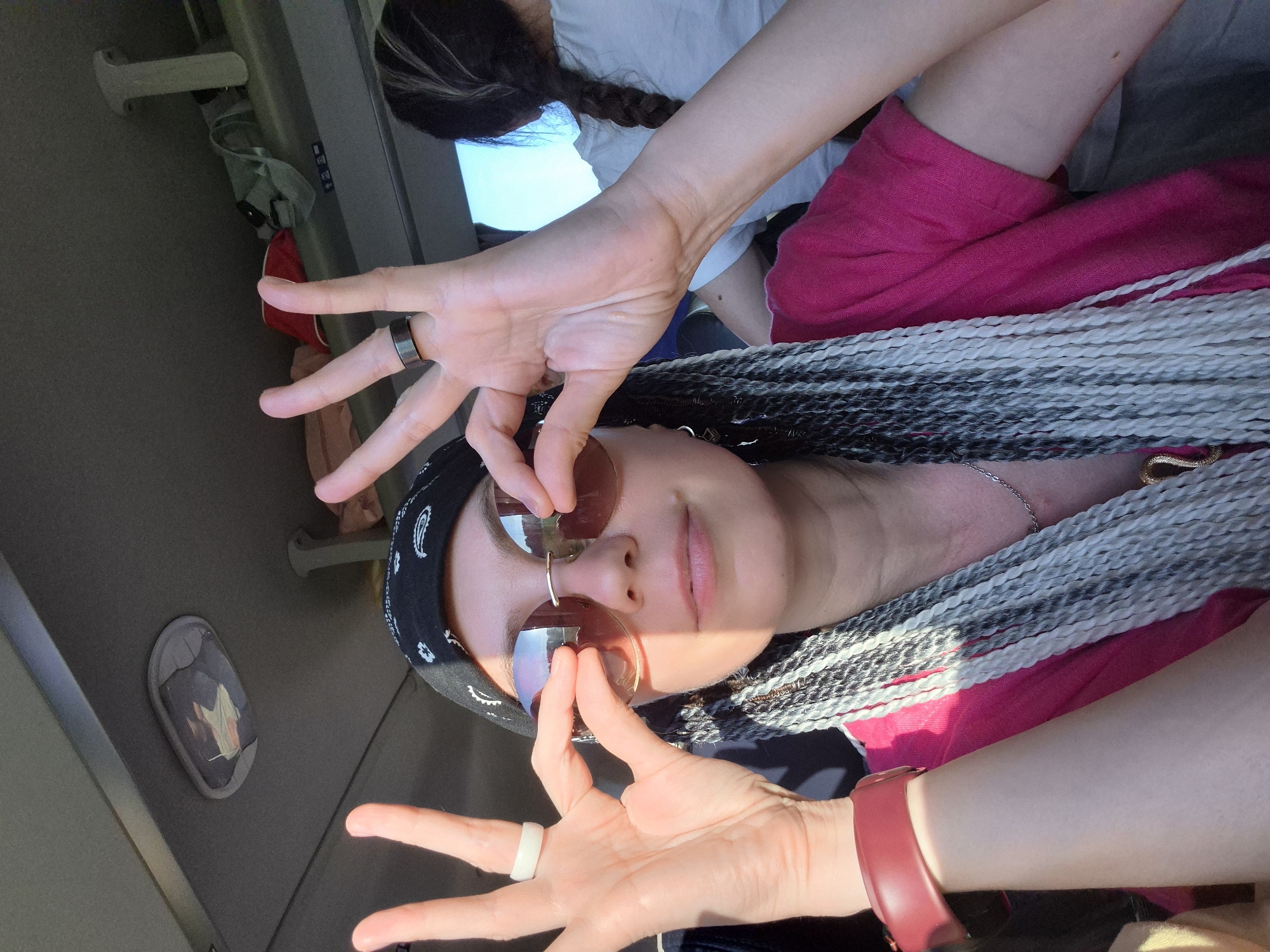 A selfie taken inside a vehicle, likely a bus, shows a woman with braided hair and sunglasses. She is wearing a pink shirt and a black bandana. Her hands are raised, and she is making a peace sign with her right hand, while her left hand is near her sunglasses. The interior of the vehicle is visible in the background, with luggage on a shelf. The lighting suggests it is daytime. The location is the city of Verkhniye Tatyshly, Russia.