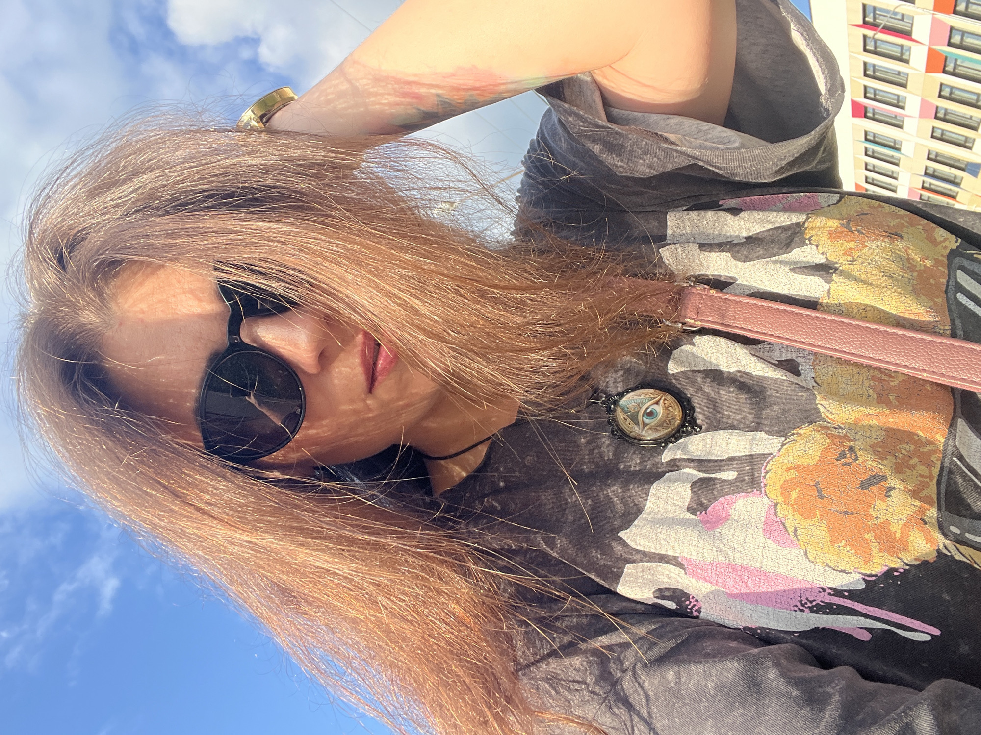 A medium shot captures a woman from the chest up, posing for a selfie outdoors. She has long, light brown hair that partially obscures her face, which is covered by dark sunglasses. A gold watch is visible on her right wrist as she touches her head. She is wearing a dark gray t-shirt with a graphic print and a pendant necklace featuring an eye design. A pink strap from a bag is visible across her chest. The background consists of a bright blue sky with scattered clouds and a modern building with colorful window frames in the distance. The lighting suggests it is daytime.