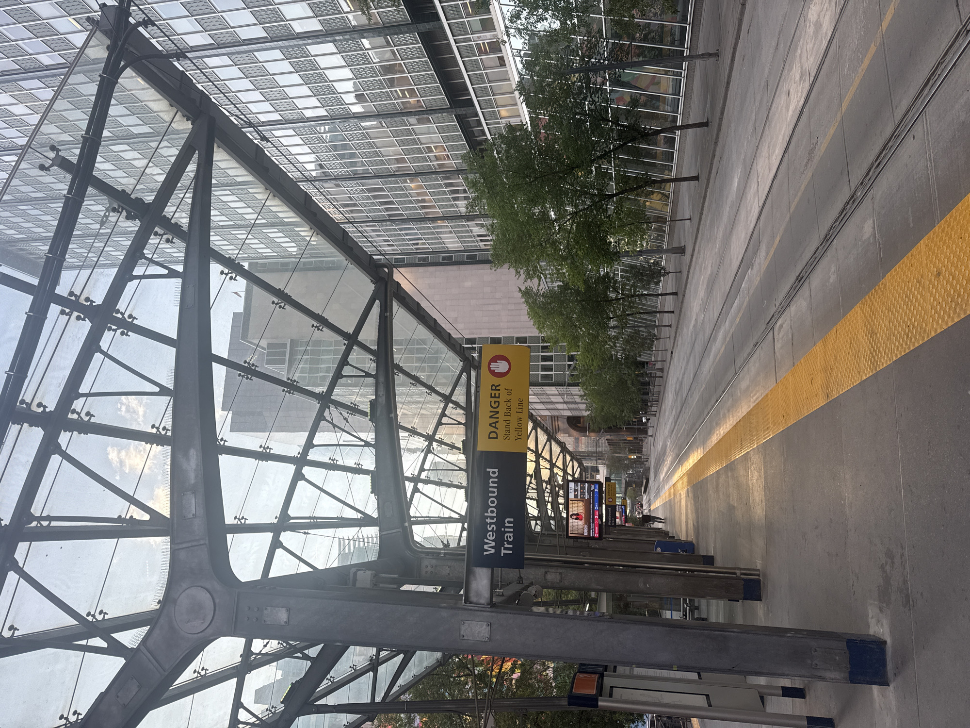 The image shows a westbound train platform in Calgary, Canada. The platform is covered by a modern glass and steel structure. To the left, a sign indicates "Westbound Train." To the right of this sign, a yellow sign displays "DANGER" and "Stand Back of Yellow Line" with a hand symbol. A single person walks away from the camera down the platform towards the train tracks. The train tracks run parallel to the platform, with a distinct yellow tactile warning strip along the edge. In the background, a row of trees lines the sidewalk, beyond which is a tall, modern building with a glass facade. The sky is visible through the glass canopy, with some clouds. The overall scene depicts a public transportation infrastructure in an urban environment.