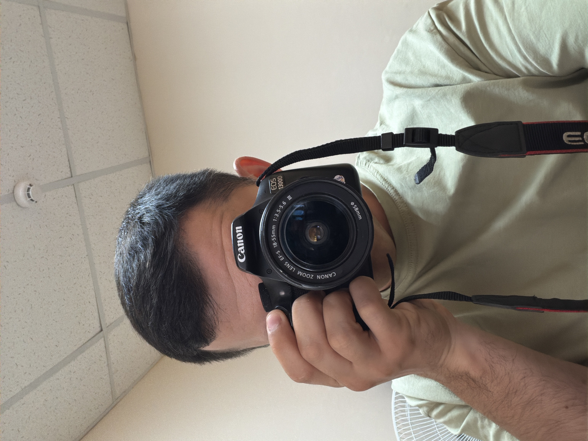 A man is holding a Canon EOS 1300D camera with a 18-55mm lens in front of his face, obscuring his eyes and nose. The camera is black with white lettering, and a black strap with red accents is attached. The man has short, dark hair, and is wearing a light green or beige t-shirt. In the background, a white, textured ceiling with a fire sprinkler is visible, along with a plain, off-white wall. A white fan is partially visible in the bottom right corner. The location of the image is not in Izhevsk, Russia.
