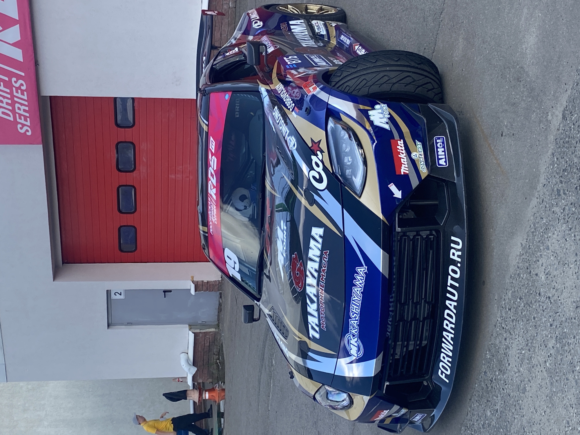 A custom-painted Nissan Z car, adorned with numerous sponsor logos and the number "19," is parked on a paved surface in front of a building with a large red industrial door. The car features a vibrant, multi-colored livery with metallic gold, blue, and white accents. Prominently displayed on the car's hood are the logos "TAKAYAMA" and "MK KASHIYYAMA." The front bumper has the website "FORWARDAUTO.RU" written in white. The car is angled, with its front wheels turned. To the left of the car, a person wearing a yellow shirt and a gray cap is walking away from the viewer. The building appears to be a garage or workshop, with a smaller gray door labeled "2" to the left of the red door. The ground is a rough, gray asphalt or concrete surface. A pink banner with "DRIFT SERIES" is visible at the top of the building. The scene is likely at a drift racing event or a related venue.