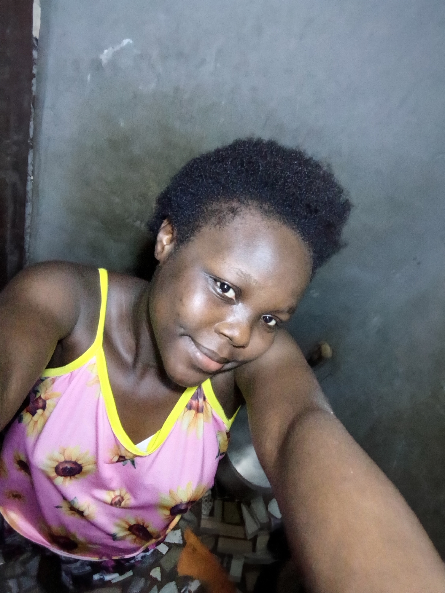 A young Black female, with dark skin and short black hair, poses for a selfie in what appears to be a dimly lit, indoor location. She is wearing a pink tank top with a sunflower print and a yellow trim. Her right arm is extended, holding the camera. Her left arm is bent, with her hand resting near her cheek. The background is a gray, textured wall. The overall lighting is low, creating shadows. Based on the provided clues, this image was taken in the city of Onitsha, Nigeria.