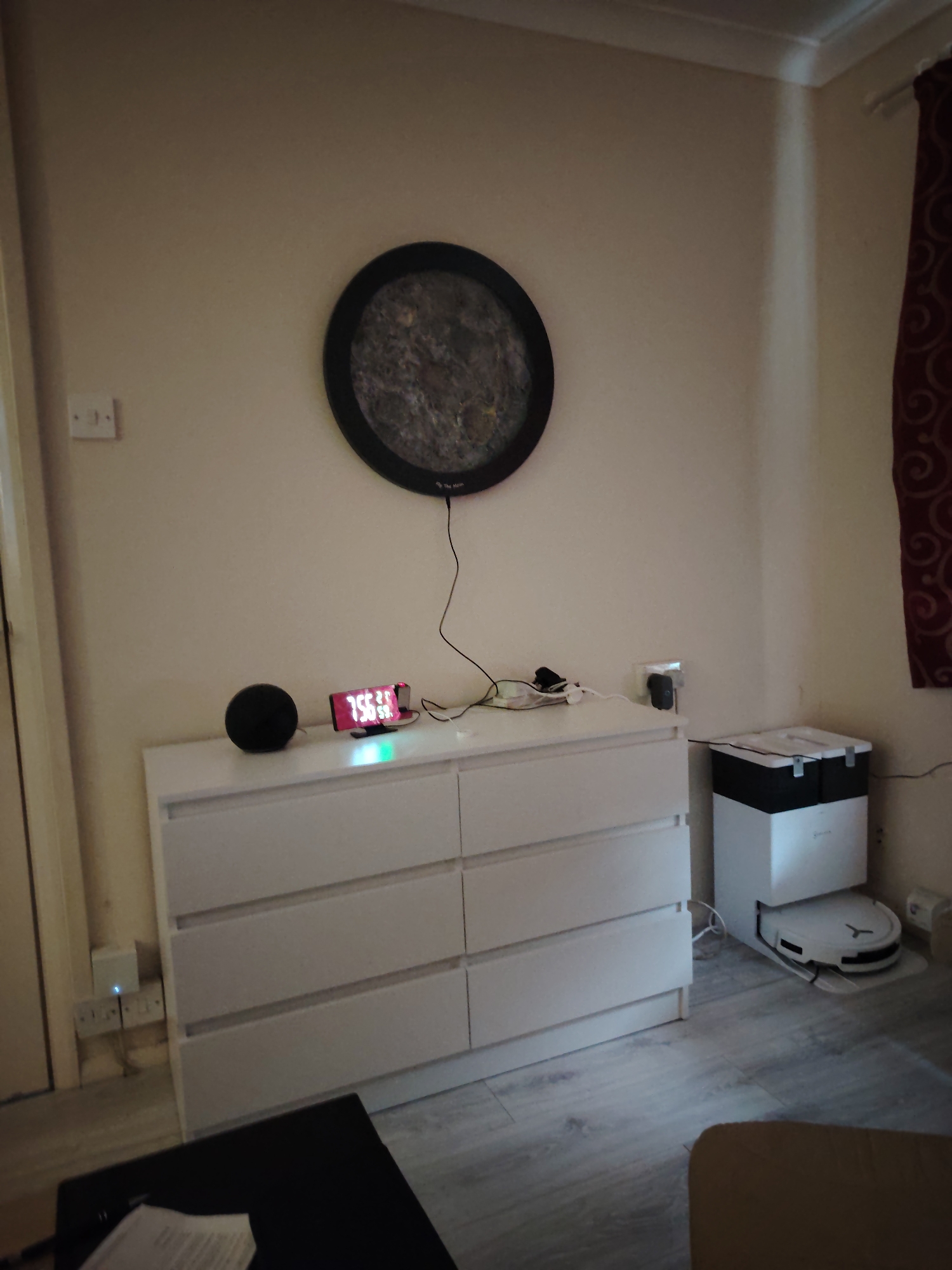 This image captures an interior scene within a residential room located in Huntingdonshire, United Kingdom. The room features light beige walls, light grey wood-effect laminate flooring, white skirting boards, and white crown molding.
A white dresser with six drawers, configured as two columns of three, is centrally positioned against the wall. On its surface, from left to right, a black spherical smart speaker is present, followed by a digital alarm clock displaying "7:56:59" in red LED numbers with an illuminated green base. Various power cables and a power adapter are also located on the dresser. Above the dresser, a black circular frame encloses a grey, textured surface, possibly depicting a lunar landscape, and is labeled "The Moon." A black power cable descends from this wall-mounted item, connecting to a power source on the dresser.
To the right of the dresser, a white UK-standard double electrical wall socket is visible, with a black plug inserted into its right outlet. Adjacent to the dresser on the lower right, a white robotic vacuum cleaner with a black top is docked at a larger white and black self-emptying or charging station. A power cable extends from this station towards the wall.
On the left side of the image, a white door frame is visible. Below it, near the floor, another white UK-standard double electrical wall socket is present, with a white power strip connected, displaying a blue indicator light. A dark, flat surface, possibly a table or laptop, with a white piece of paper, is partially visible in the bottom left foreground. A section of a light brown upholstered item, likely furniture, is visible in the bottom right foreground. A red curtain with an elaborate swirling pattern is visible on the far right side of the room. No individuals are present in the scene.