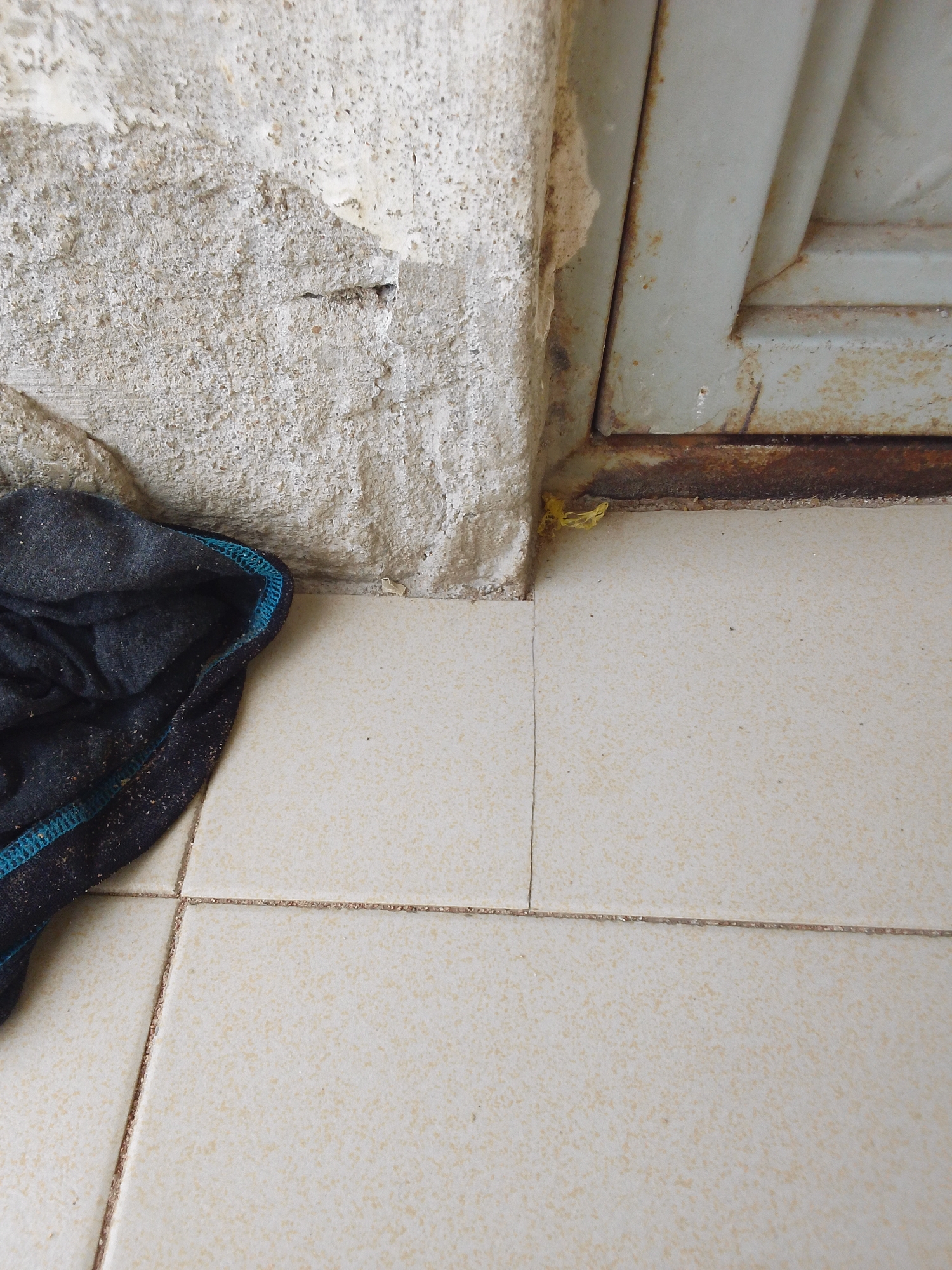 The image depicts a close-up view of a corner where a rough, textured wall meets a tiled floor and a metal doorframe. A dark blue and black cloth, possibly a rag or a piece of clothing, is crumpled on the left side of the frame, resting on the light-colored tiled floor. The wall shows signs of wear and tear, with chipped plaster revealing a rougher texture underneath. The metal doorframe appears to be aged, with visible rust along the bottom edge. A small piece of bright yellow material, possibly plastic debris, is stuck in the corner where the wall, doorframe, and floor converge. The floor is composed of square tiles with a speckled pattern, laid with dark grout lines that form a grid. The lighting suggests it is daytime, likely indoors or in a shaded outdoor area, given the lack of harsh shadows. The scene is one of everyday neglect or disuse, with no specific activity or event clearly taking place. The environment seems to be utilitarian, possibly a back entrance, storage area, or a less-maintained part of a building in Jalingo, Nigeria. There is no visible text in the image.