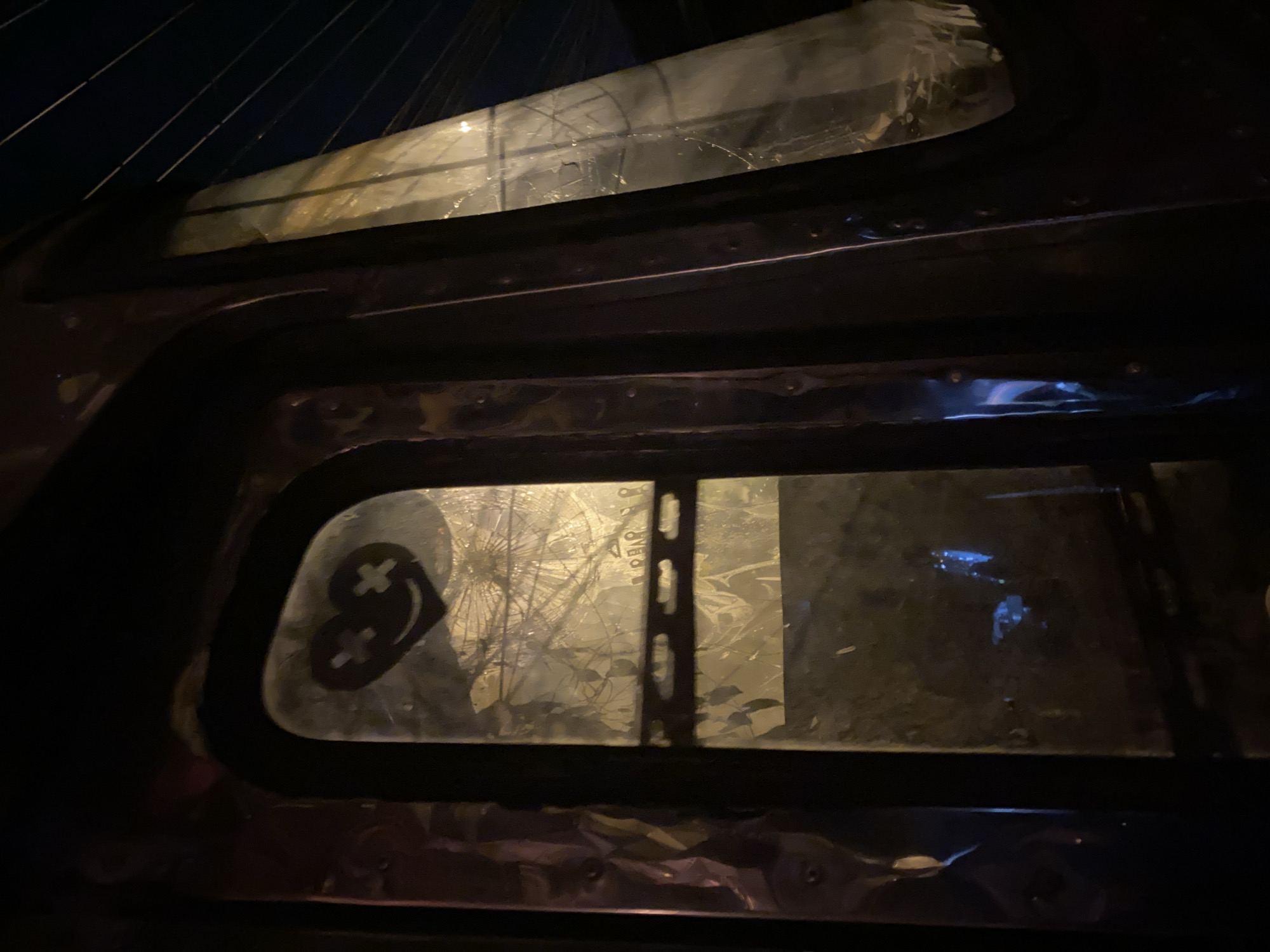 The image shows the interior of a vehicle, specifically the rear area, with a window visible. A heart-shaped sticker with X's for eyes is on the window. Through the window, there are reflections and visual obstructions, possibly indicating an outside scene at night. The reflections make it difficult to discern any clear objects outside the vehicle. It is very dark inside and outside the vehicle. Based on the location "ตำบลเกาะพะงัน, Thailand," the scene is likely in a rural or coastal area, but without further detail, it is difficult to provide more information about the specific location. The cables visible through the top window could indicate that the vehicle is in a location where powerlines or suspension cables (like those of a bridge) are present.