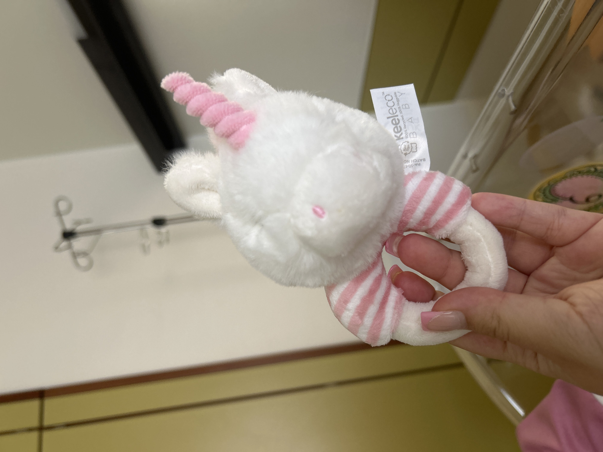 In a hospital room, likely in Gummersbach, Germany, a person's hand with light-colored skin and french-manicure style nails holds a white plush unicorn rattle. The rattle has a pink horn, pink stripes on its body, and a white ring. A white tag is attached to the rattle with the brand name "keeleco" and the words "100% Recycled, 100% Huggable BABY". In the blurred background, an IV pole and a medical bassinet can be seen.
