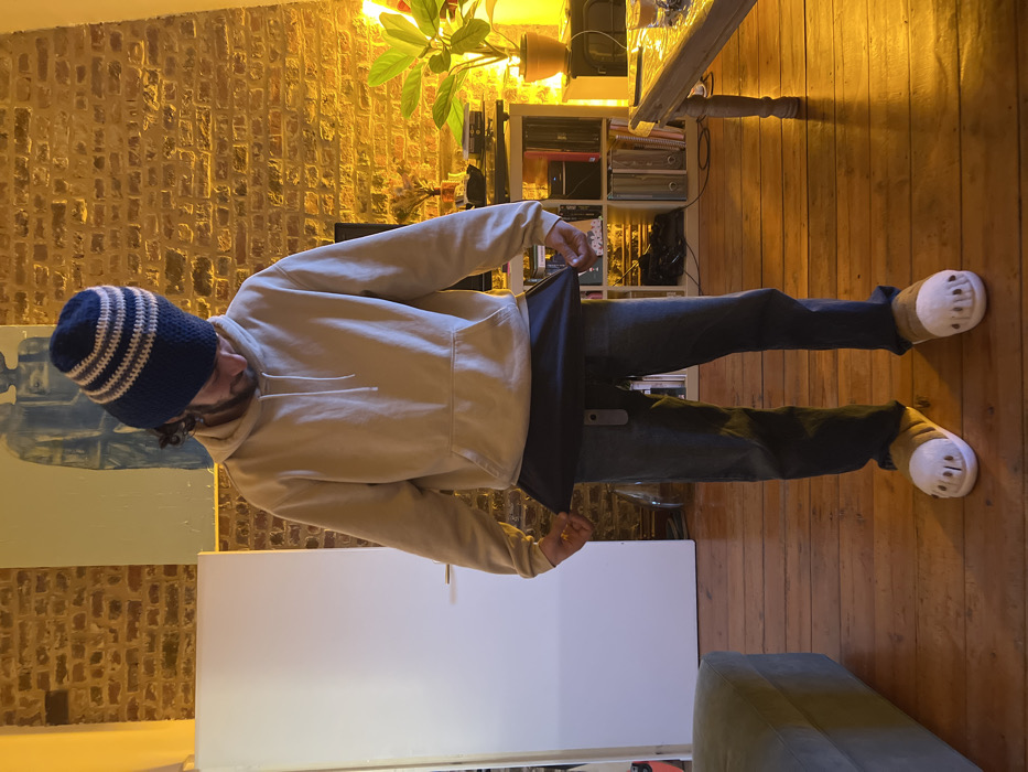 The image displays an indoor scene featuring a person standing in what appears to be a living space or room. The person is an adult male with a beard and dark, curly hair, mostly covered by a knitted beanie hat. The hat is horizontally striped in alternating shades of blue and off-white/cream. He is wearing a light beige or tan long-sleeved hoodie with a front pouch pocket and dark blue jeans. On his feet, he wears distinctive white and beige slipper-like shoes that have a unique, almost claw-like or cartoonish bear paw design at the front.
The individual is standing with his body slightly angled, looking down and to his right. He is holding a dark, rectangular object, possibly a laptop, folder, or book, with both hands at waist level. The object is slightly open or angled away from the viewer.
The background consists of a warm, inviting setting. To the right of the person, there is an exposed brick wall, illuminated by a warm, yellowish light emanating from an unseen source above and to the right, suggesting evening or artificial lighting. In the mid-ground, a wooden table with a plant and other items is visible, and below it, a white shelving unit filled with various books and objects can be seen. The flooring is made of warm-toned wooden planks. To the person's left, a light-colored wall features a large, light blue and white abstract painting or canvas. Further to the left, a large white rectangular object, possibly a refrigerator or a cabinet, is visible. In the bottom left corner of the frame, a portion of a green or olive-colored upholstered sofa or armchair is present.
The overall atmosphere suggested by the warm lighting and indoor setting is cozy and relaxed. No discernible text is visible in the image.