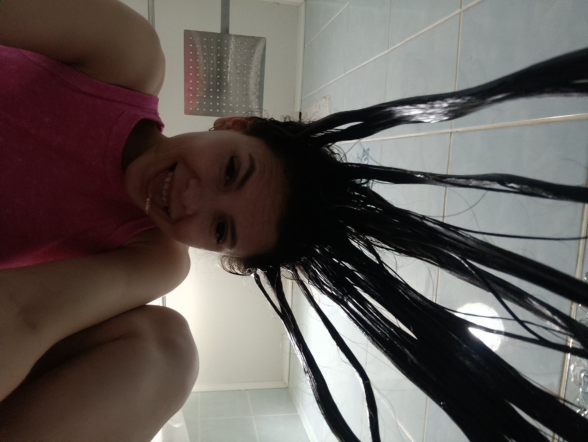 A woman is positioned in a bathroom setting, displaying her wet, dark hair towards the camera. She is wearing a pink tank top. A shower head is visible on the wall. The bathroom features light blue tiles. The exact location is unspecified, in Unknown, Unknown.