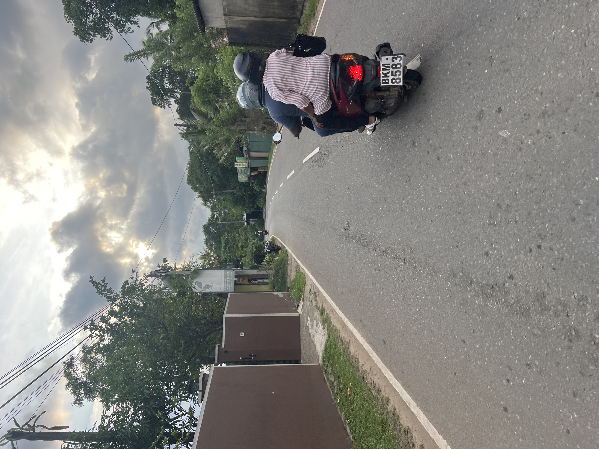 Angulugaha Junction, Sri Lanka: An eye-level, slightly rotated image depicts two people on a moped traveling on a gray asphalt road. The driver wears a gray helmet and a dark blue shirt. The passenger behind wears a red and white striped shirt. The moped's license plate reads "BKM-8583." The sky is cloudy, with the sun peeking through. The road is bordered by green vegetation on the left and a brown-painted wall. Trees and utility poles are visible in the background.