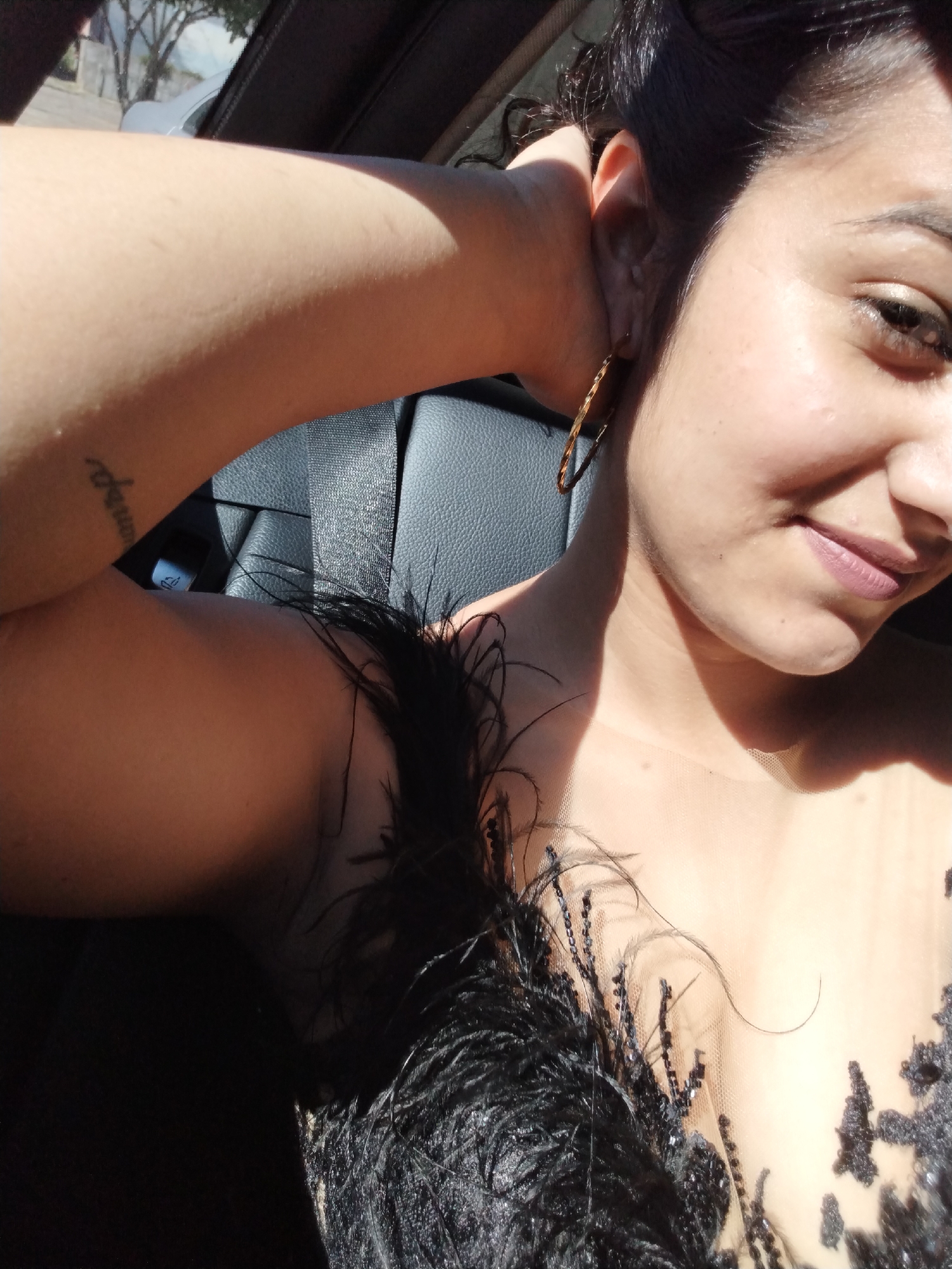 A young woman, with dark hair and brown eyes, is captured from the chest up, inside a vehicle. She is looking towards the camera with a slight smile, her head tilted back and her left arm bent, with her hand resting behind her neck. She wears a delicate, sheer black top adorned with feathers and sequins. A gold hoop earring is visible in her left ear. Sunlight streams in from the side, casting a shadow across her face and arm. The background shows the textured gray leather of a car seat and glimpses of the outside through a car window, suggesting an urban or suburban setting. Her arm has a small tattoo.