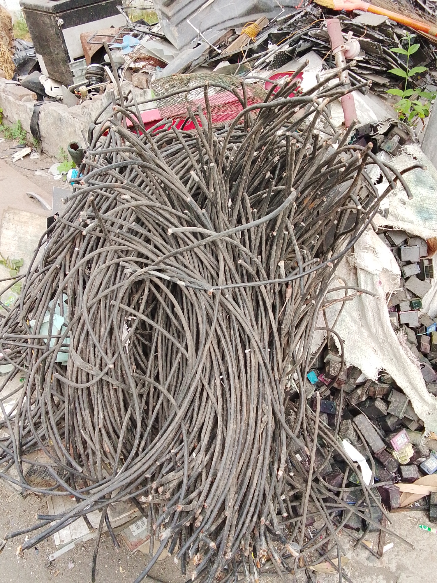 In a cluttered junkyard, likely in Bulurejo, Indonesia, a large pile of thick, black electrical cables dominates the foreground. These cables, cut and bundled together, are the primary subject. Behind the cables, the environment is a chaotic assortment of discarded materials. This includes various electronic components, fragmented plastic casings in shades of black, purple, and pink, and some white fabric. In the background, more debris is visible, including what appears to be parts of larger appliances or machinery, and a green plant growing amidst the waste. The overall impression is one of a waste disposal or recycling area, filled with an accumulation of broken and discarded items.