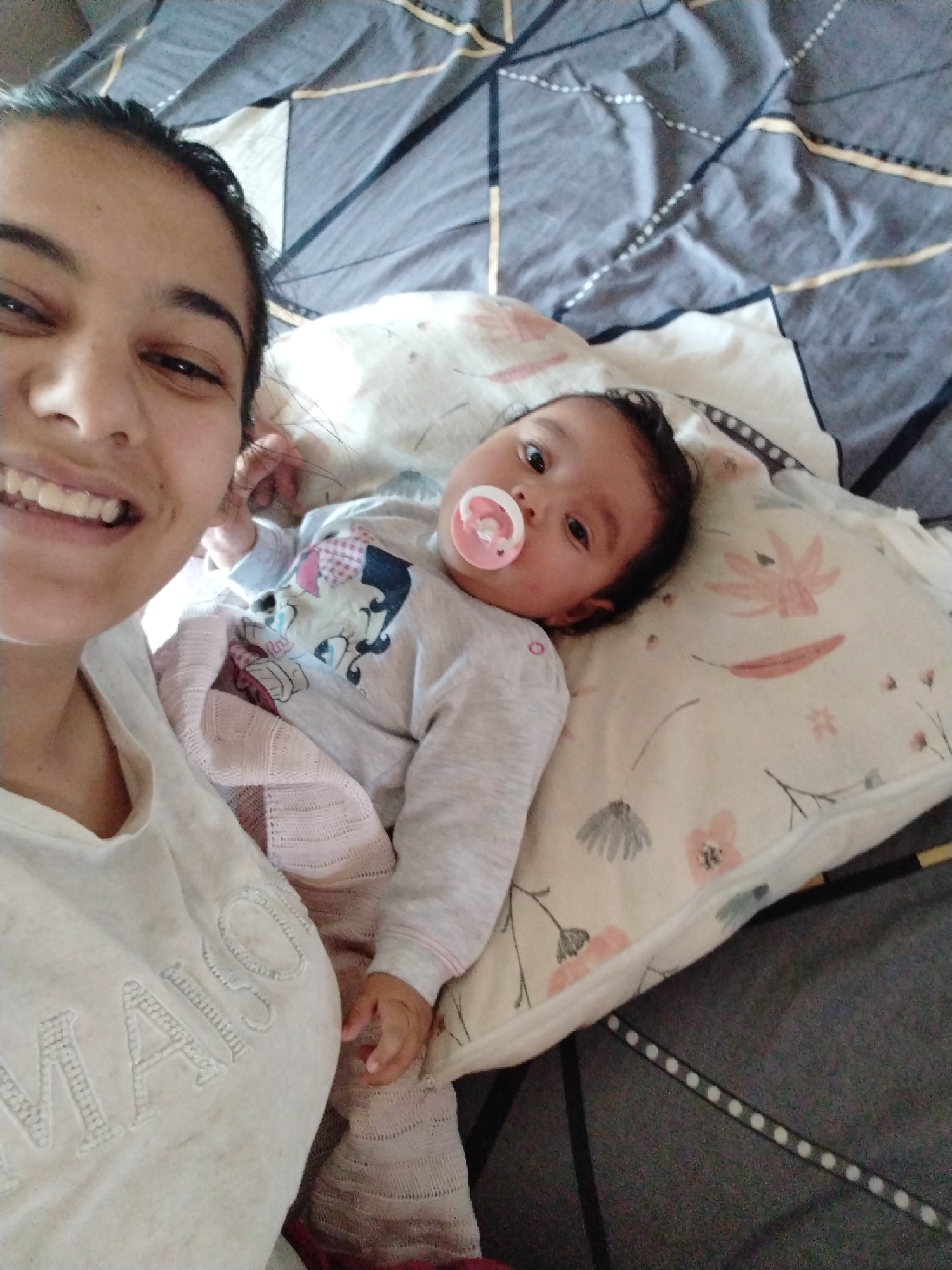 A woman and a baby are lying on a bed. The woman is on the left side of the image, smiling at the camera. She is wearing a light gray shirt. The baby is lying on a floral patterned pillow to the right of the woman, with a pink pacifier in its mouth. The baby is wearing a gray long-sleeved shirt with a graphic design on the front and light pink patterned leggings. A light pink blanket is draped over the baby. The bed is covered with a gray and white patterned duvet. The location is Unknown, Unknown.