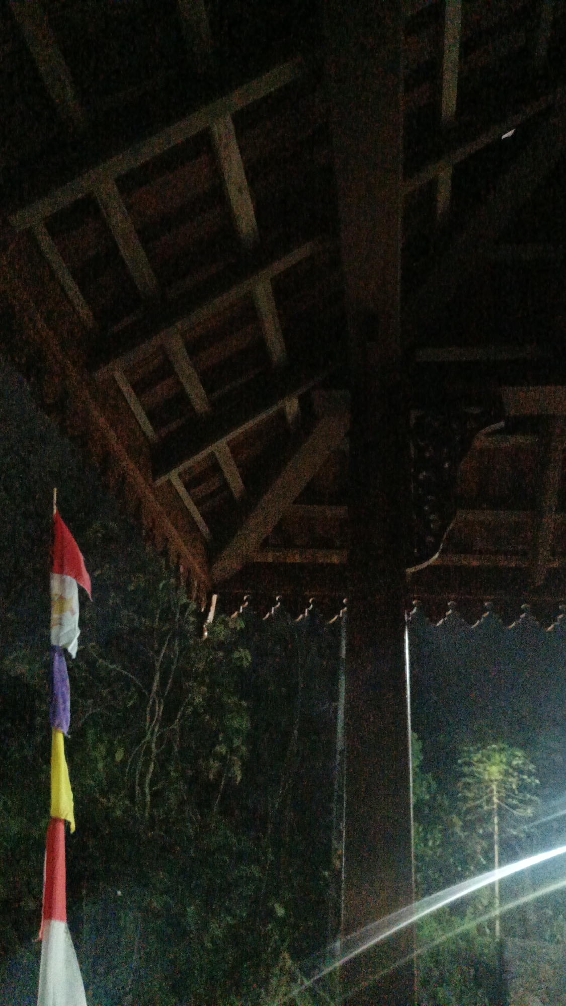 The image shows a section of a wooden gazebo roof at night in Sambi, Indonesia. The roof is constructed with dark wooden beams and planks, some of which are intricately carved. A tall pole with several decorative flags hanging from it is positioned to the left of the gazebo. The flags are colorful, with red, white, yellow, and purple sections visible. Beyond the gazebo and flags, lush green foliage and trees are discernible in the dimly lit background. The lighting suggests an outdoor setting, possibly a garden or park, with artificial lights creating reflections on a surface on the right side of the frame.