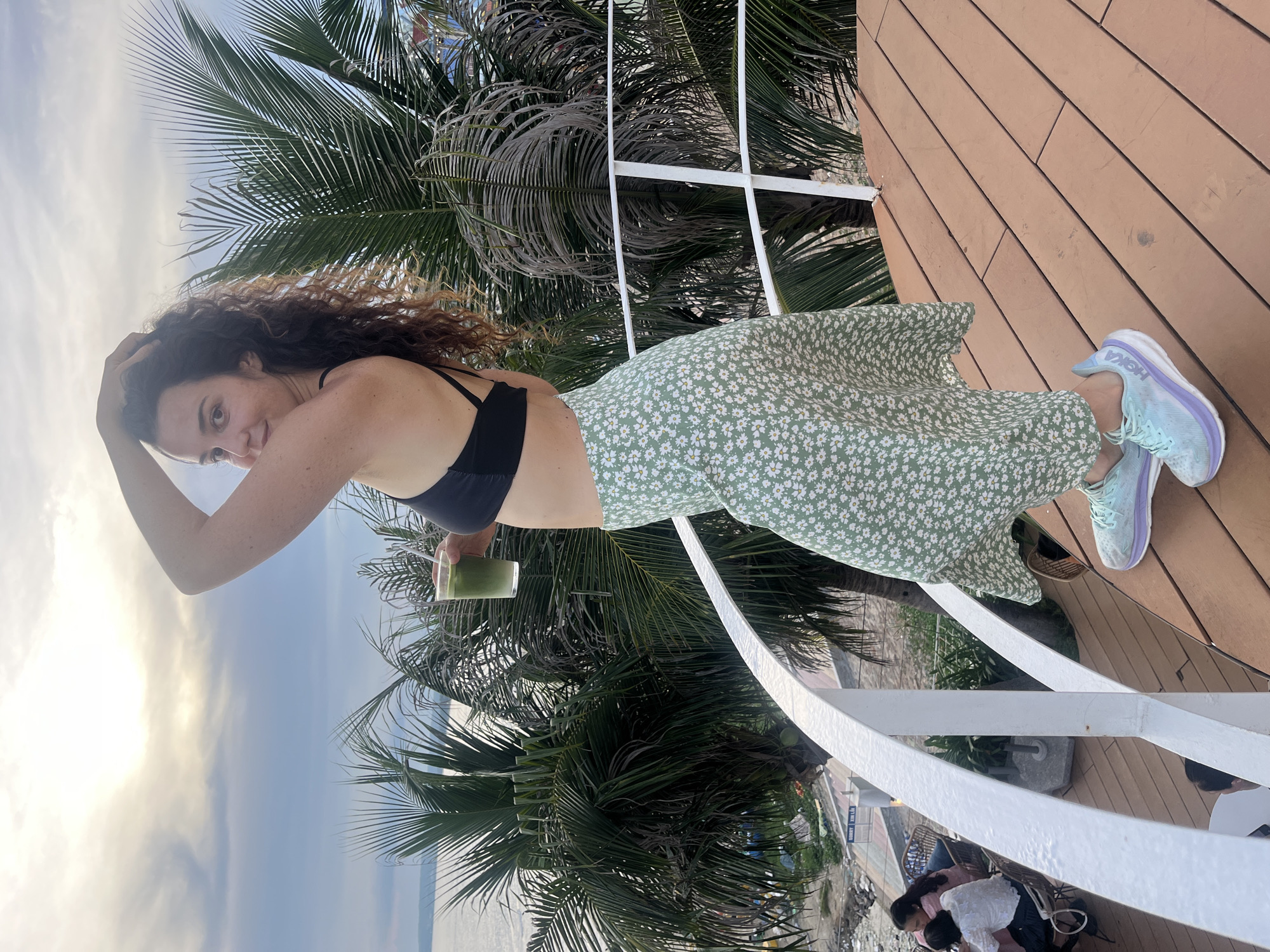 A woman with curly brown hair stands on a wooden deck overlooking a body of water and palm trees. She is wearing a black bikini top, a light green floral midi skirt, and light blue and purple sneakers. She holds a clear plastic cup with a green drink and a straw in her right hand and has her left arm raised, touching her hair. In the background, there are several palm trees and people sitting at tables. The sky is cloudy. The location is Mui Ne, Vietnam.