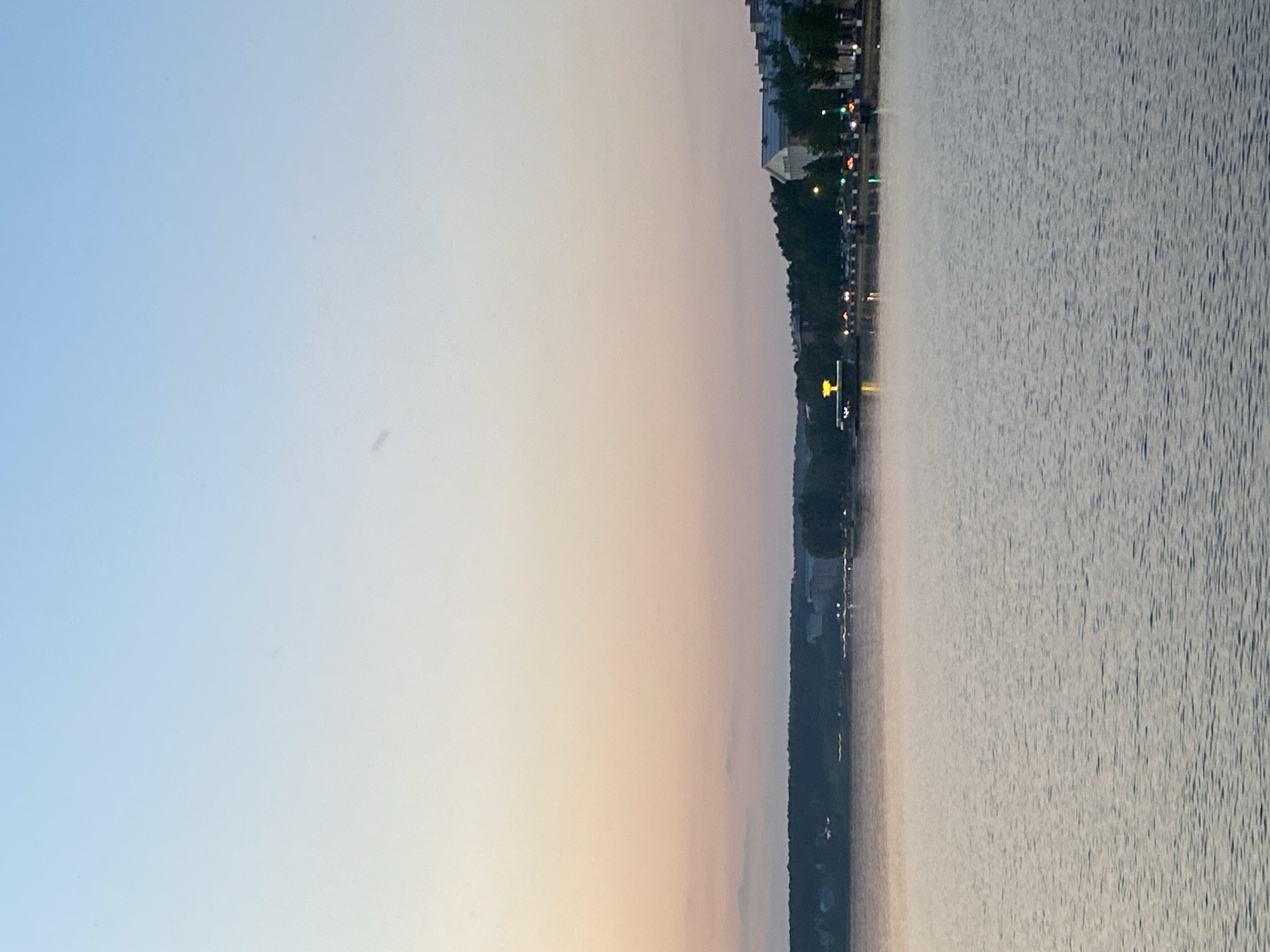 The image depicts a body of water, likely a lake or river, with a shoreline visible in the middle. The sky takes up the left side of the image, transitioning from a light blue to a peach color, indicating either sunrise or sunset. The water surface is textured with small waves, reflecting the light from the sky. Along the shoreline, there is evidence of vegetation and possibly some buildings or structures, as well as lights. Based on the information given, the location is Votkinsk, Russia. The lights along the shoreline suggest a park, road, or other developed area with the presence of infrastructure.