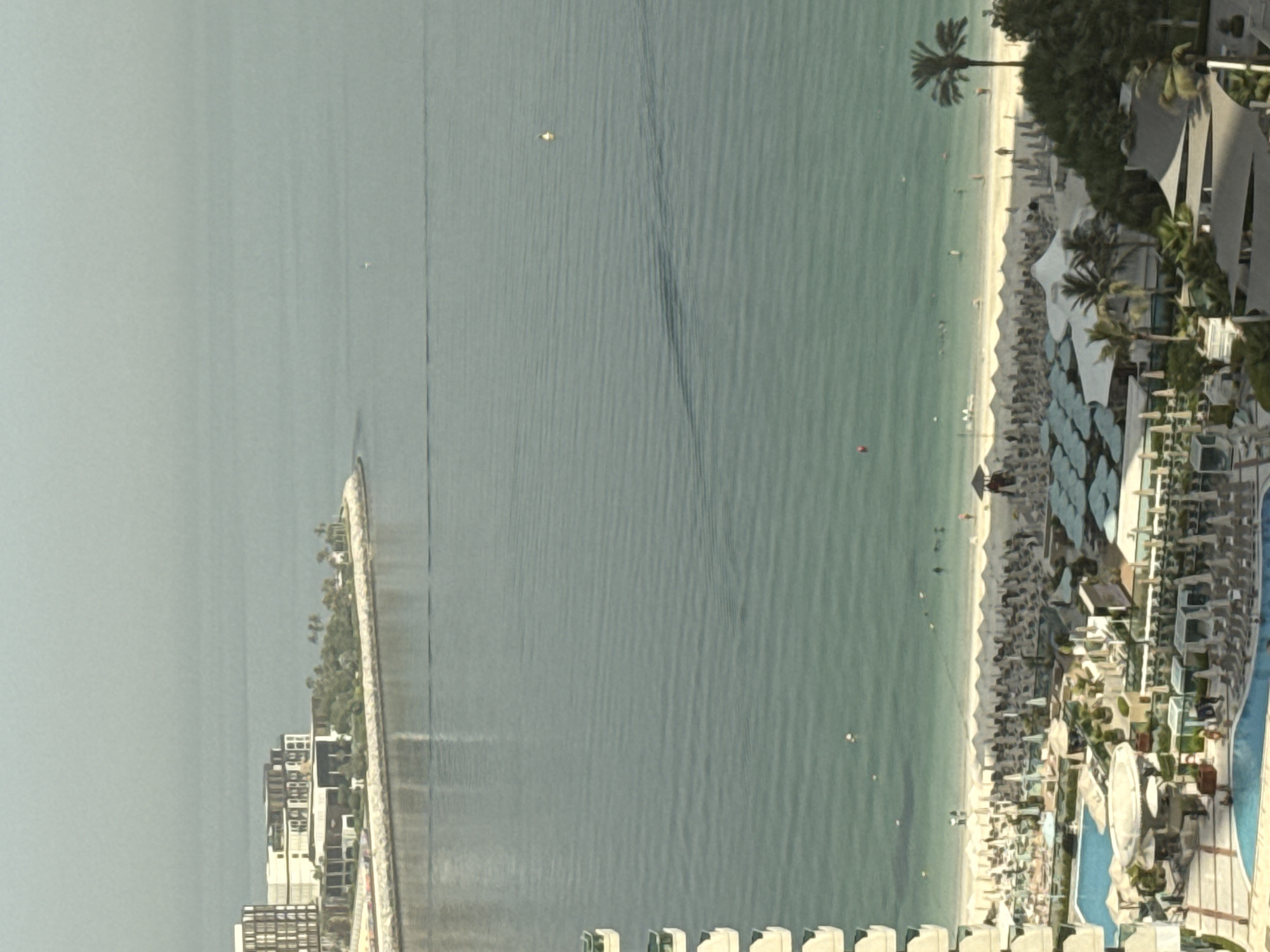 This is an aerial photograph of a developed coastline in Dubai, United Arab Emirates. In the upper left portion of the image, a modern, multi-story building complex with a dark facade and white accents is situated on a peninsula. Adjacent to this complex, a man-made rock barrier extends into the turquoise water, creating a sheltered cove. The majority of the image is dominated by the sea, with a visible transition from deeper blue offshore to a lighter, clearer hue near the shore. Gentle ripples are present on the water's surface, and two buoys are visible in the distance.
The foreground of the image features a densely populated beach resort area. A wide stretch of sand is lined with numerous white beach umbrellas and loungers. Several individuals are scattered across the beach and in the shallow water, some appearing to be swimming or wading. A resort structure with a dark roof and white supports is visible on the right side of the beach. Inland from the beach, a resort complex with multiple swimming pools, cabanas, and shaded seating areas is depicted. Lush green trees, including palm trees, are interspersed throughout the resort grounds. The scene suggests a recreational and tourist-oriented environment.