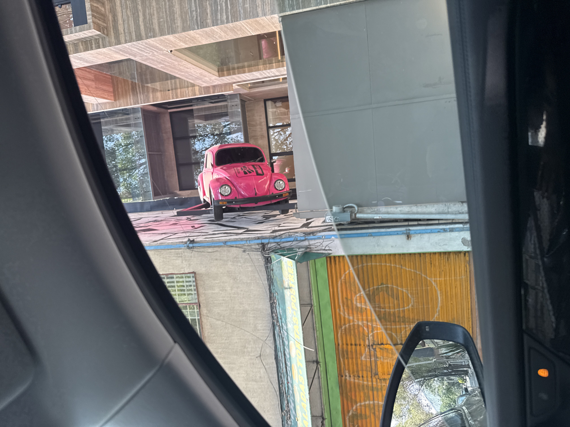 This media file captures a street scene in Mexico City, viewed from inside a vehicle, likely through a car window. The main subject is a vibrant pink Volkswagen Beetle, prominently displayed on what appears to be a concrete platform or a lower roof level of a modern building. The car features black text on its hood, which reads "KABOM" with a stylized explosion graphic.
The immediate background behind the pink Beetle includes reflections of trees and sky in large glass windows of the building it's situated on. The building itself has textured, possibly wooden or stone-clad, facades and a contemporary architectural style with overhangs and recessed sections.
Below the level of the pink car, the scene transitions to older, more traditional urban architecture. There's a light-colored building with a barred window. Further down, a long green and blue sign is visible, which appears to be a business sign. Partial text on this sign can be deciphered as "ENSALADAS TOSTADAS POSTRES," indicating a food establishment offering salads, tostadas, and desserts. Next to this sign, a corrugated metal roll-up door, painted bright orange-yellow with abstract white graffiti, is visible.
The bottom right of the image shows a partial view of a car's side mirror, reflecting more of the urban environment and trees. An orange indicator light is also visible on the interior trim of the viewer's car. The overall impression is a sunny day, with bright light and clear visibility. The image has a somewhat disorienting quality due to the perspective from inside a moving or parked car, capturing multiple layers of urban environment and reflections.