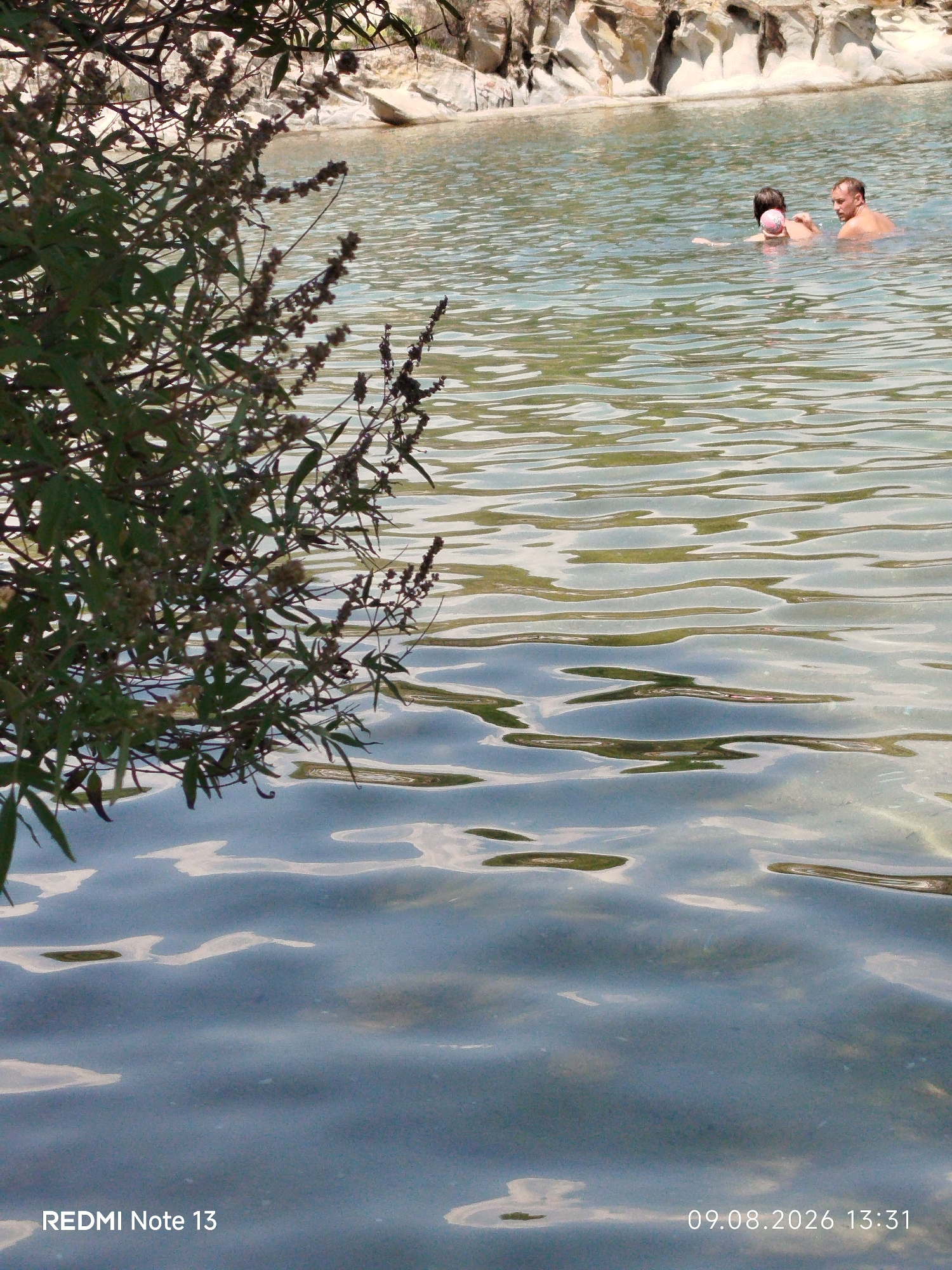 The image shows a natural body of water, likely a lake or a calm sea, with a rocky shoreline in the background. The water is clear and has visible ripples reflecting the sunlight. In the middle distance, two individuals are swimming. One is a man, and the other is a person with dark hair, possibly a woman or a child, holding what appears to be a pink object. To the left of the frame, there is a dense cluster of green foliage with small, dried flower heads, partially obscuring the view of the water and the background. The foreground is dominated by the rippling surface of the water. The timestamp "09.08.2026 13:31" and "REDMI Note 13" are visible in the bottom corners of the image, suggesting it was taken with a Redmi Note 13 phone on August 9, 2026, at 13:31. The location is identified as Sithonia Municipal Unit, Greece.