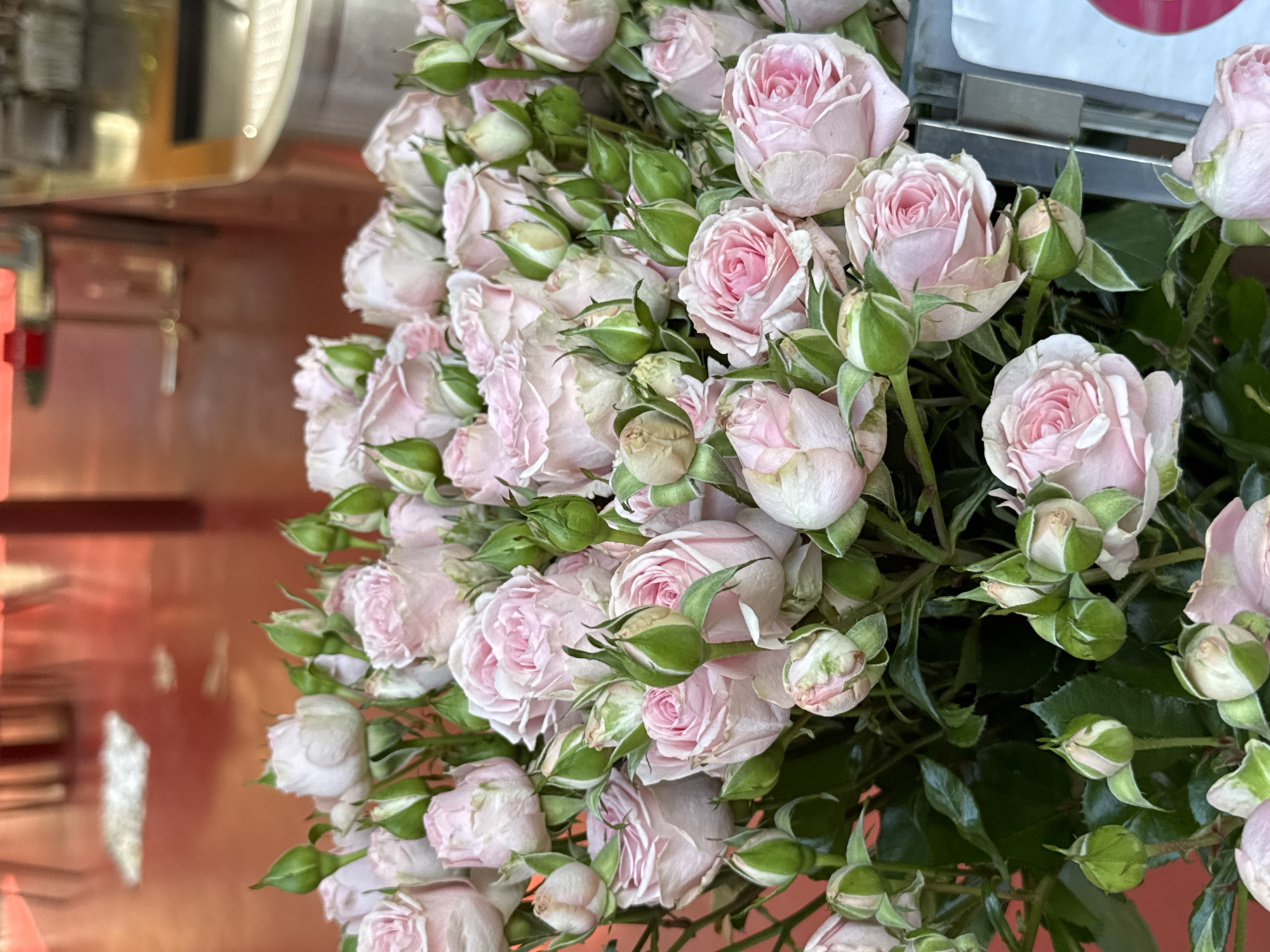 A close-up shot shows a bouquet of light pink roses with green buds and leaves. The flowers are tightly packed, filling most of the frame. The background includes blurred elements of an interior with a reddish-brown surface and some metal structures. The location is Yerevan, Armenia.