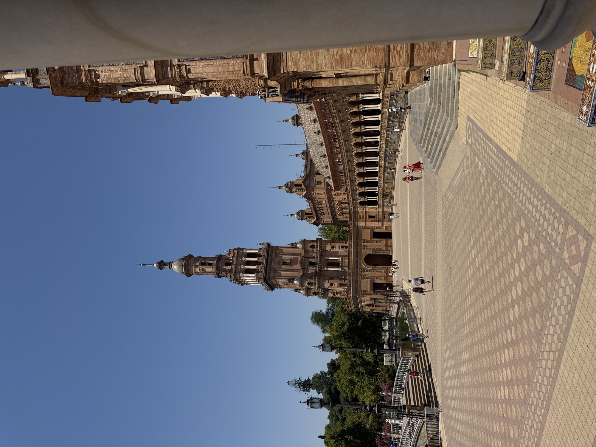 The image shows a bright, sunny day with a clear blue sky. The scene is an outdoor plaza with intricate architecture and tiled pathways. In the foreground, a wide expanse of light-colored tiles creates a patterned surface. Several people are visible walking on the plaza.
Dominating the background is a grand, ornate building with multiple towers and arches, constructed from reddish-brown brick. The building features extensive decorative elements, including columns and what appear to be tiled roofs. A tall, slender tower with a dome-like structure rises prominently from the building.
To the left of the building, a section of what seems to be a decorative railing or balustrade is visible, with trees and lampposts behind it. Further to the left, a part of another building, possibly a colonnade or walkway, is seen, also made of brick and featuring decorative stonework and white columns.
The image captures a lively public space, with people engaging in leisurely activities like walking. The overall atmosphere appears relaxed and pleasant, indicative of a tourist destination. The architecture suggests a historical or culturally significant location. Based on the provided location context, this is likely Plaza de España in Seville, Spain, known for its unique architecture and sprawling plaza.