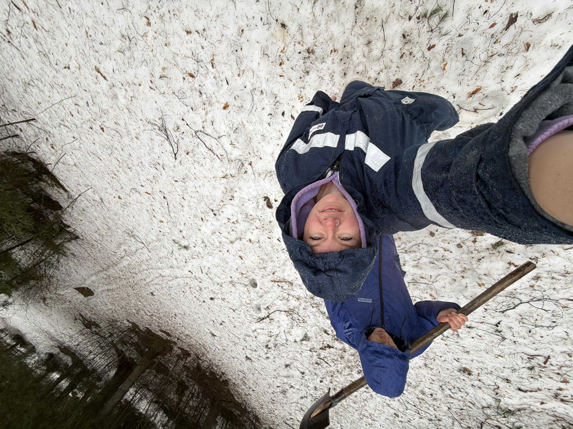 A top-down perspective shows a person lying in the snow, possibly in a forest. The person is wearing a dark blue hooded jacket over a purple hooded garment and is holding a shovel and a wooden stick. The background is a snowy ground covered with twigs and fallen leaves, transitioning to a dense forest area on the left side of the image. The location is possibly near Chaikovsky, Russia.