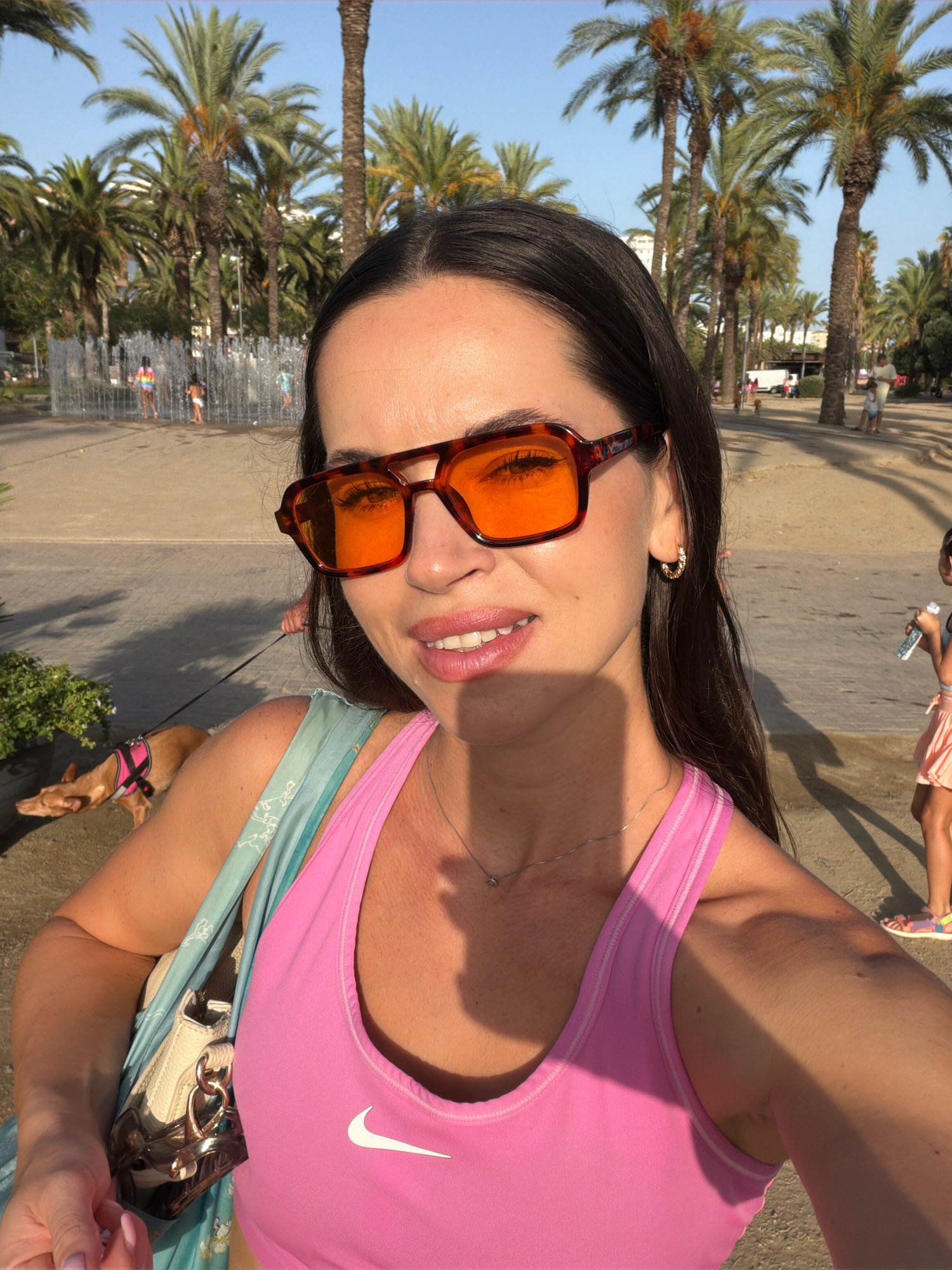 In Salou, Spain, a woman wearing orange-tinted square sunglasses and a pink Nike tank top takes a selfie outdoors. She has long dark hair and a delicate necklace. To her left, a light-colored handbag hangs from her shoulder, partially obscured by a light blue patterned scarf. A small dog on a leash, wearing a pink harness, walks in the background. In the distance, children play near a fountain, and palm trees line a sandy path. The scene suggests a sunny day in a park or beachfront area.